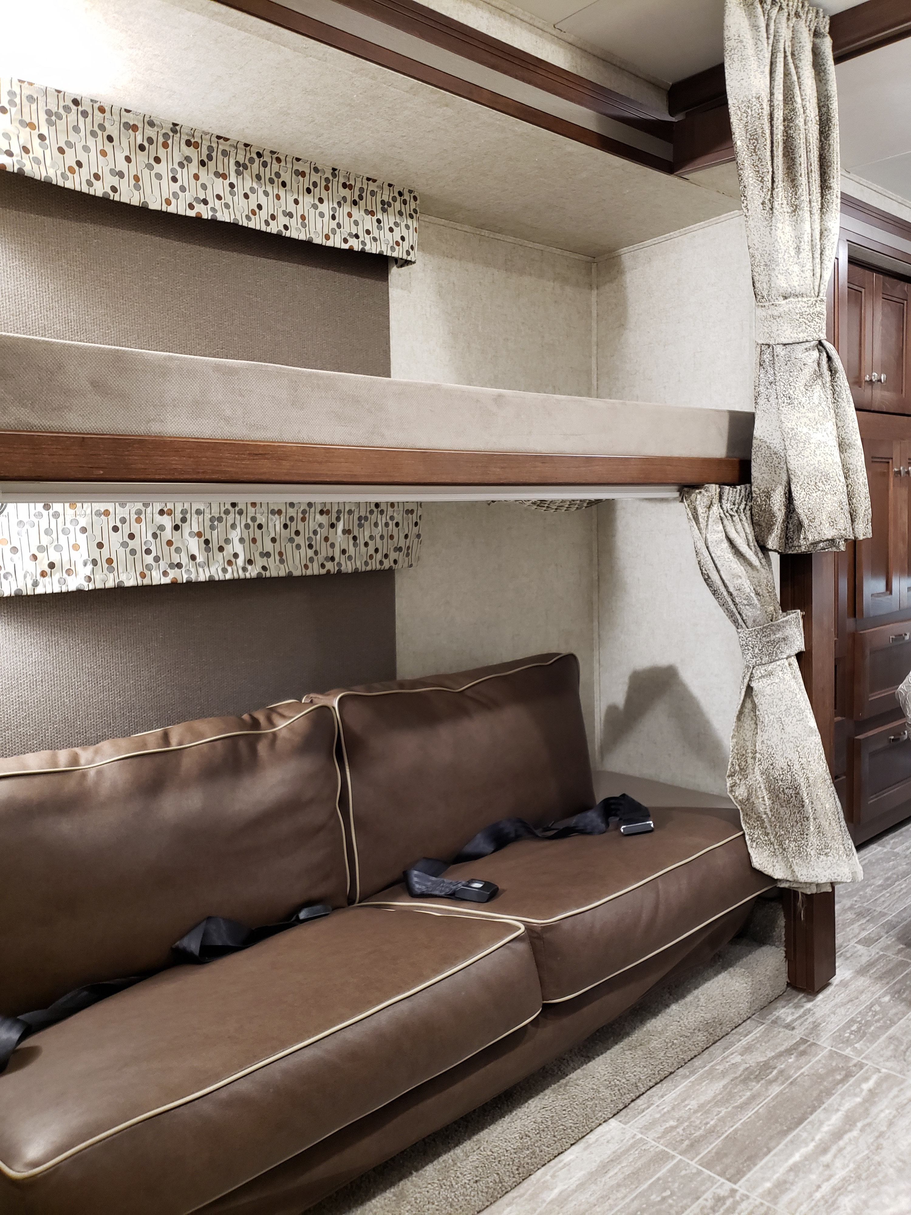 Compact RV rental interior featuring a bunk bed over a brown leather couch with seatbelts and patterned valances.. Other Other 2019