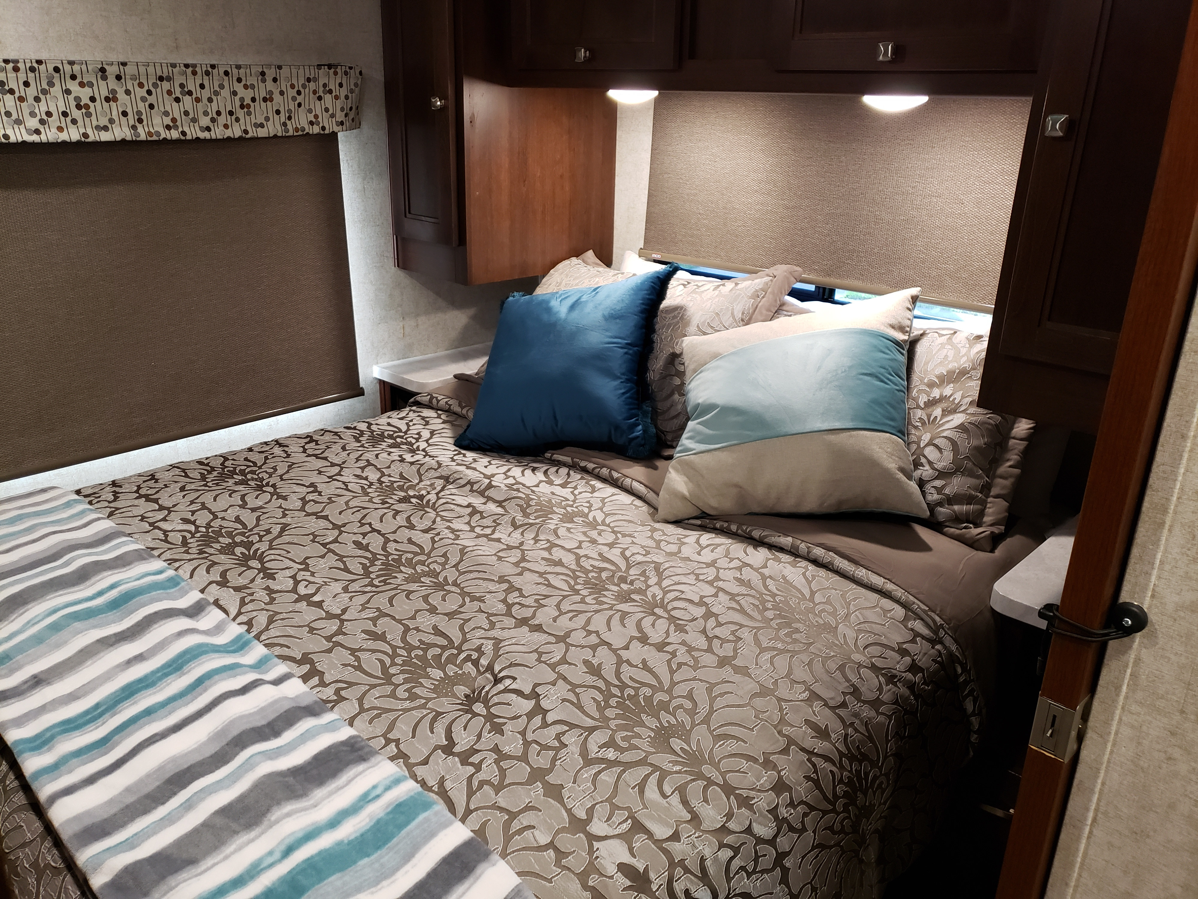 Cozy rv rental bedroom showcasing a patterned bed, blue accent pillows, brown window blinds, and overhead storage.. Other Other 2019