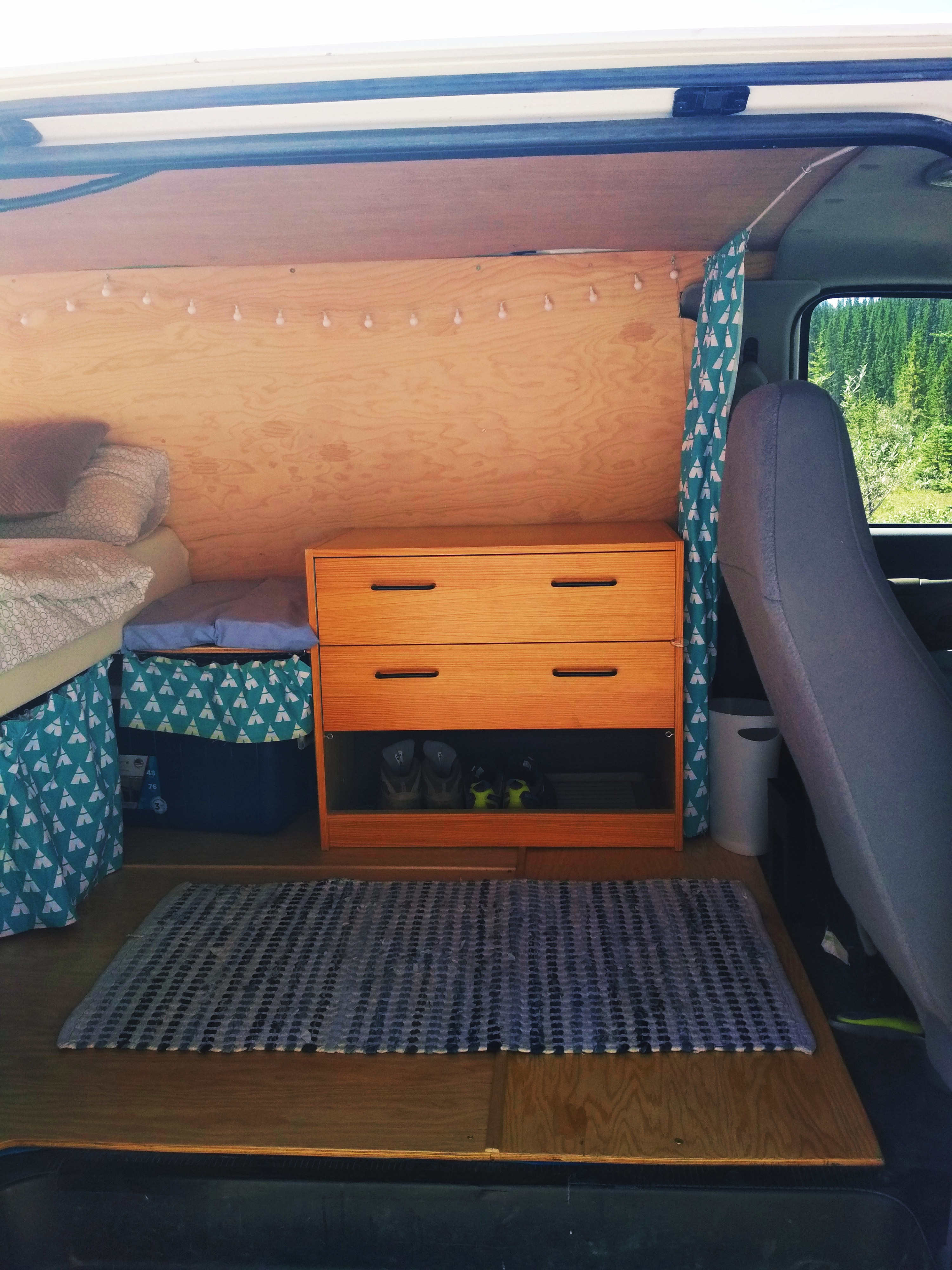 Inside a cozy RV rental van: custom plywood interior with bed, dresser, storage, patterned curtain, and forest views.. Ford Econoline 2006
