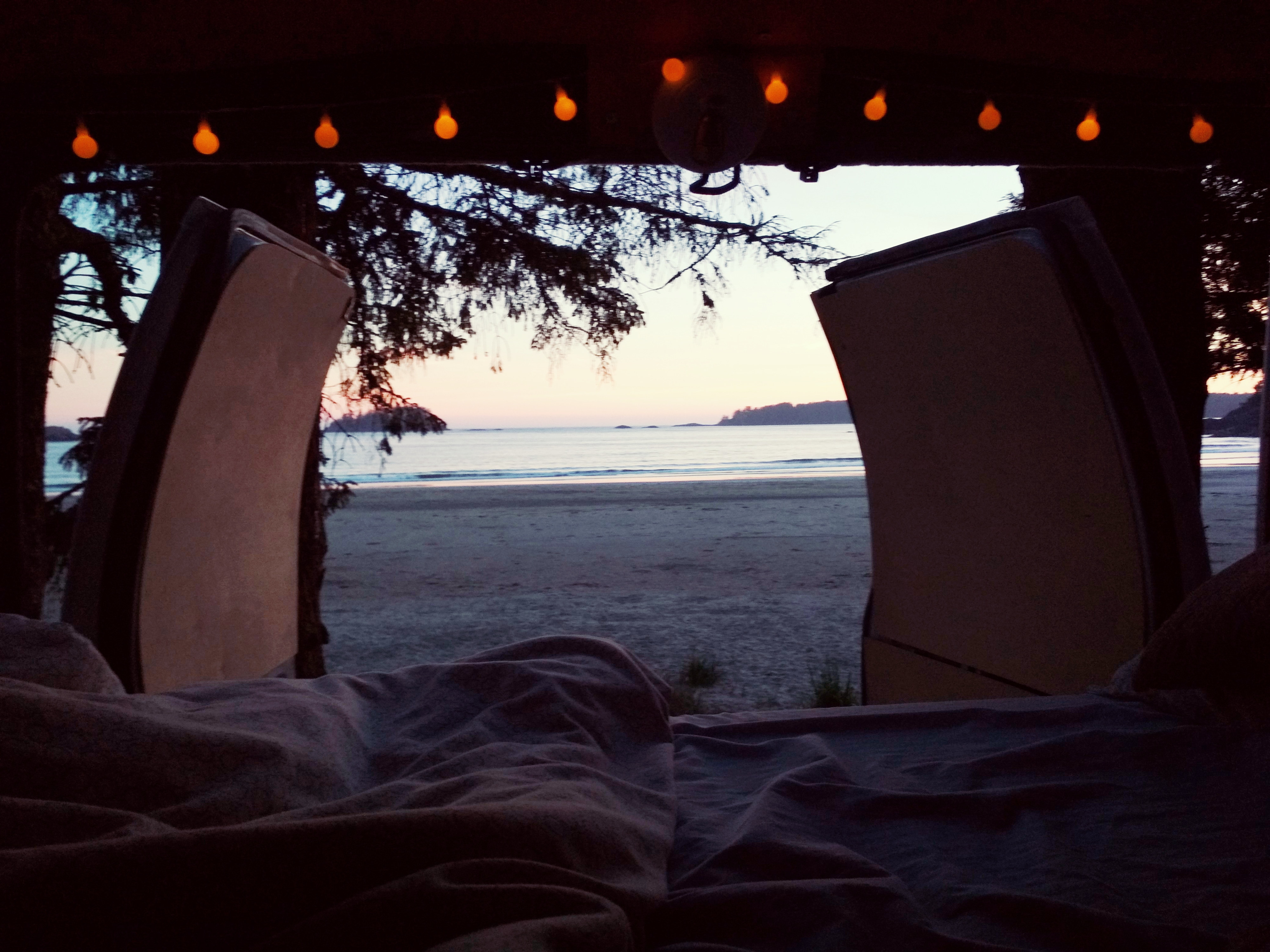 Cozy rv rental bed view of a serene beach sunset. String lights glow inside the van, framing the tranquil ocean and distant shore.. Ford Econoline 2006