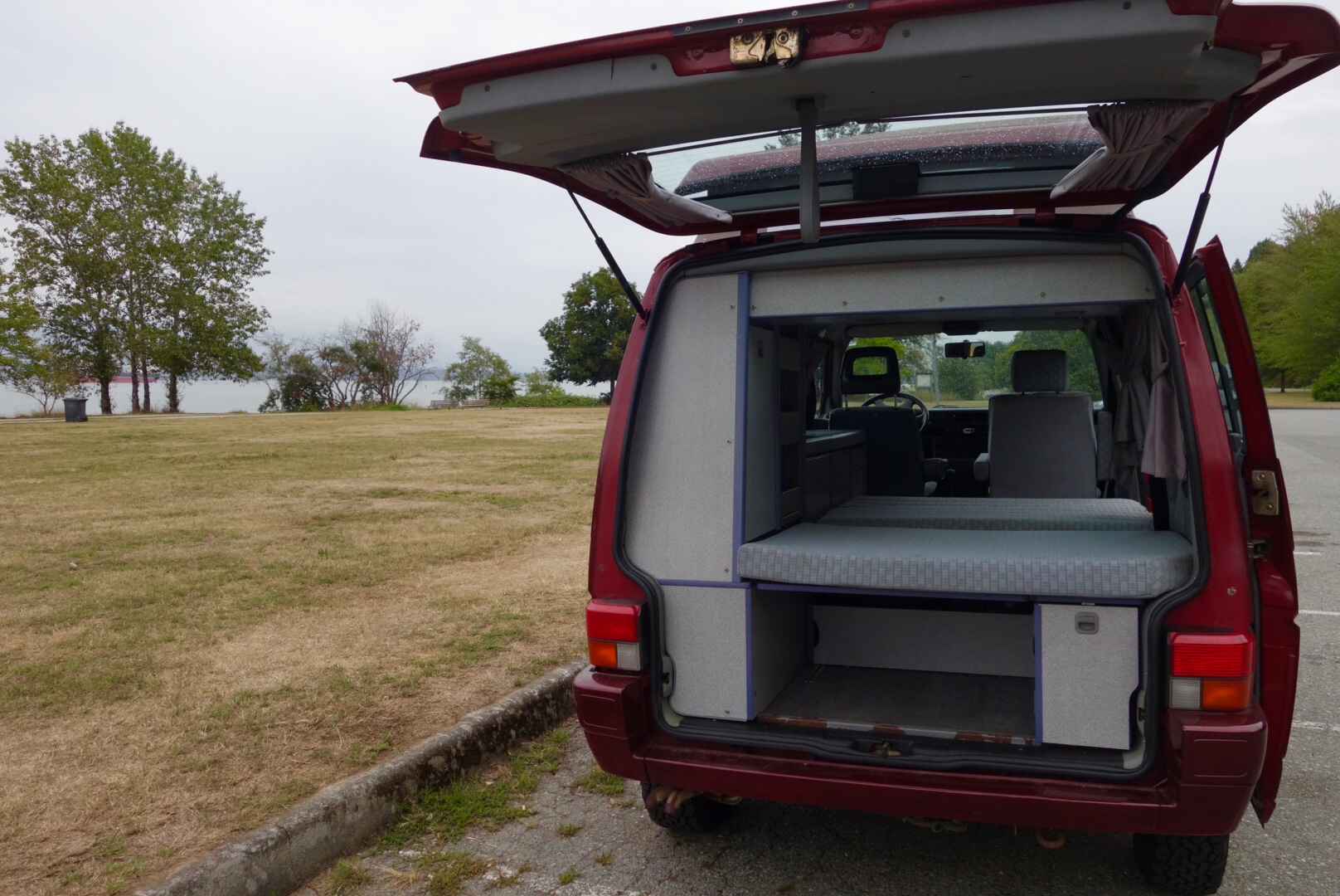 RV rental: Red camper van with tailgate open, showing comfortable bed & interior. Parked by a scenic grassy field and bay.. Volkswagen T4 Westfalia 1992