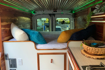 Step inside this RV rental! Cozy camper van interior with a bed, pillows, wood paneling, and green overhead lighting.. Ford Transit 2016
