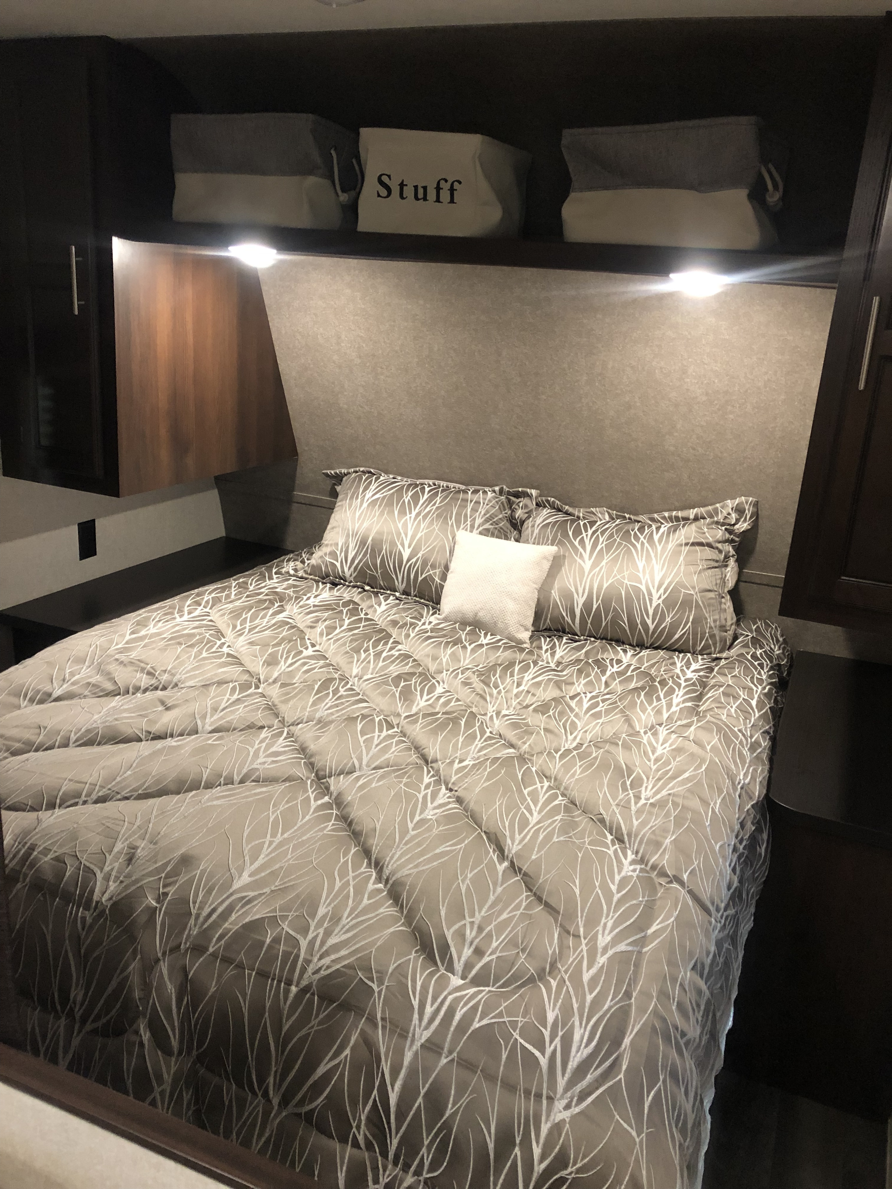 Cozy RV rental bedroom featuring a grey bedspread with white branch patterns, pillows, and overhead storage bins, one labeled "Stuff.". Jayco Flight 2019