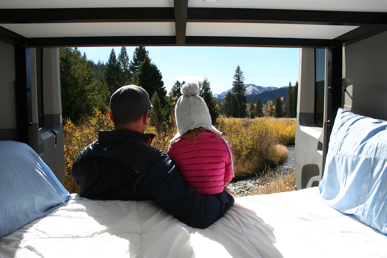 Inside their RV rental, a father and child gaze at a stunning mountain, forest, and river view. Perfect road trip stop.. Ford Transit 350 2019