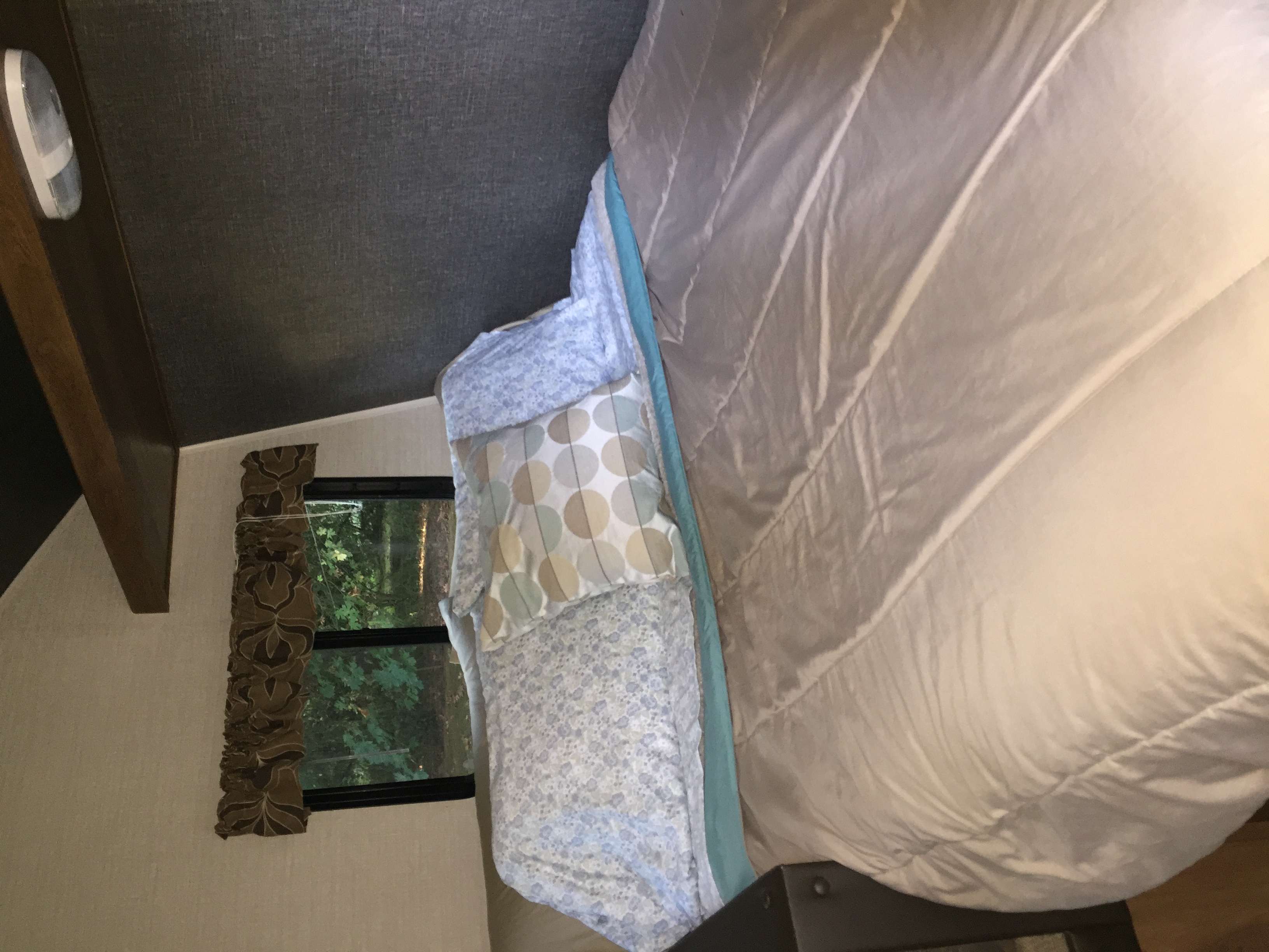 Inside a comfortable RV rental bedroom, featuring a bed with patterned pillows and a window overlooking lush green nature.. Keystone RV Summerland Mini 2015