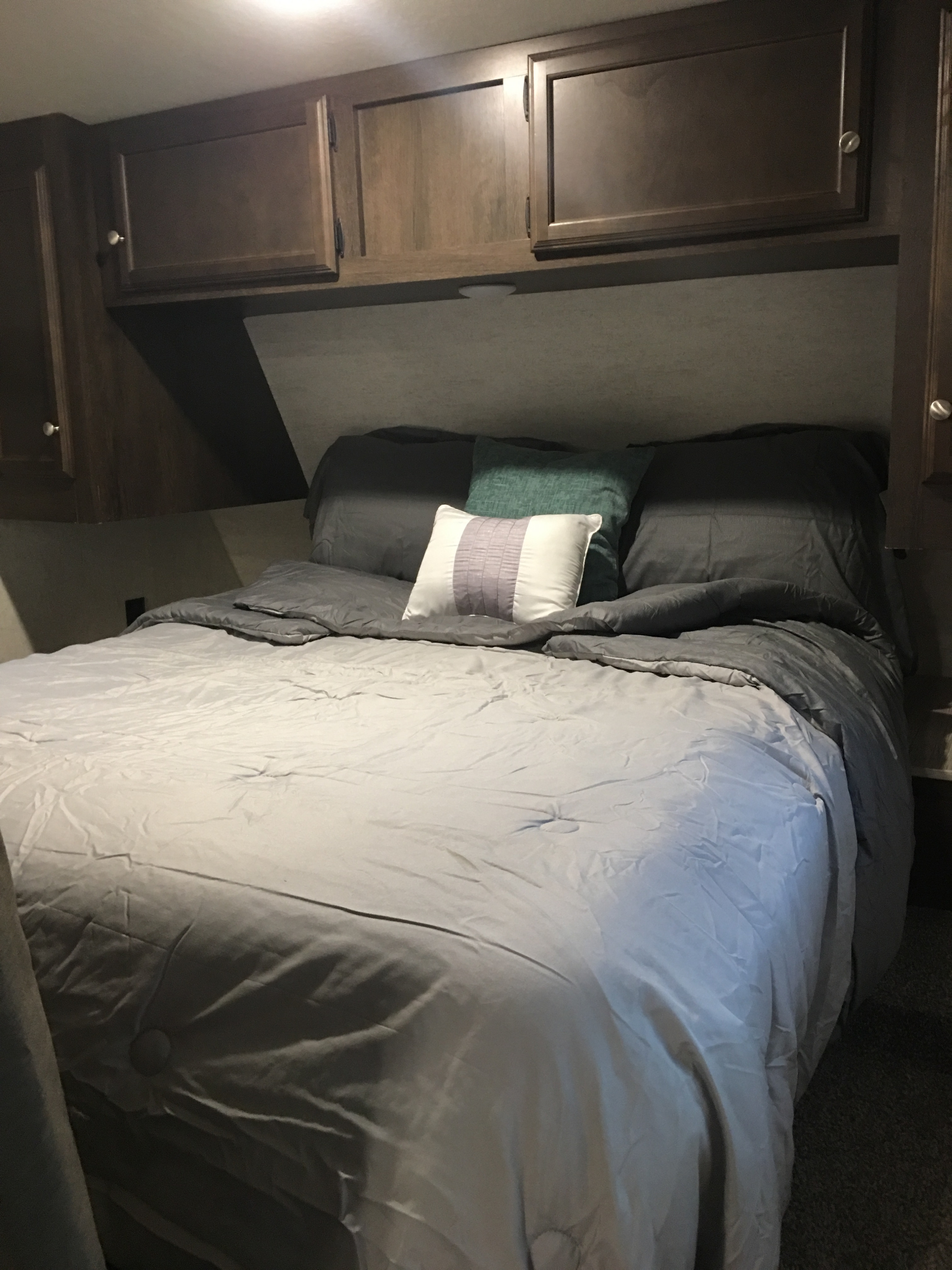 Compact RV rental bedroom: comfy bed with grey bedding, accent pillows, and dark wood overhead cabinets.. Starcraft Autumn Ridge 2018