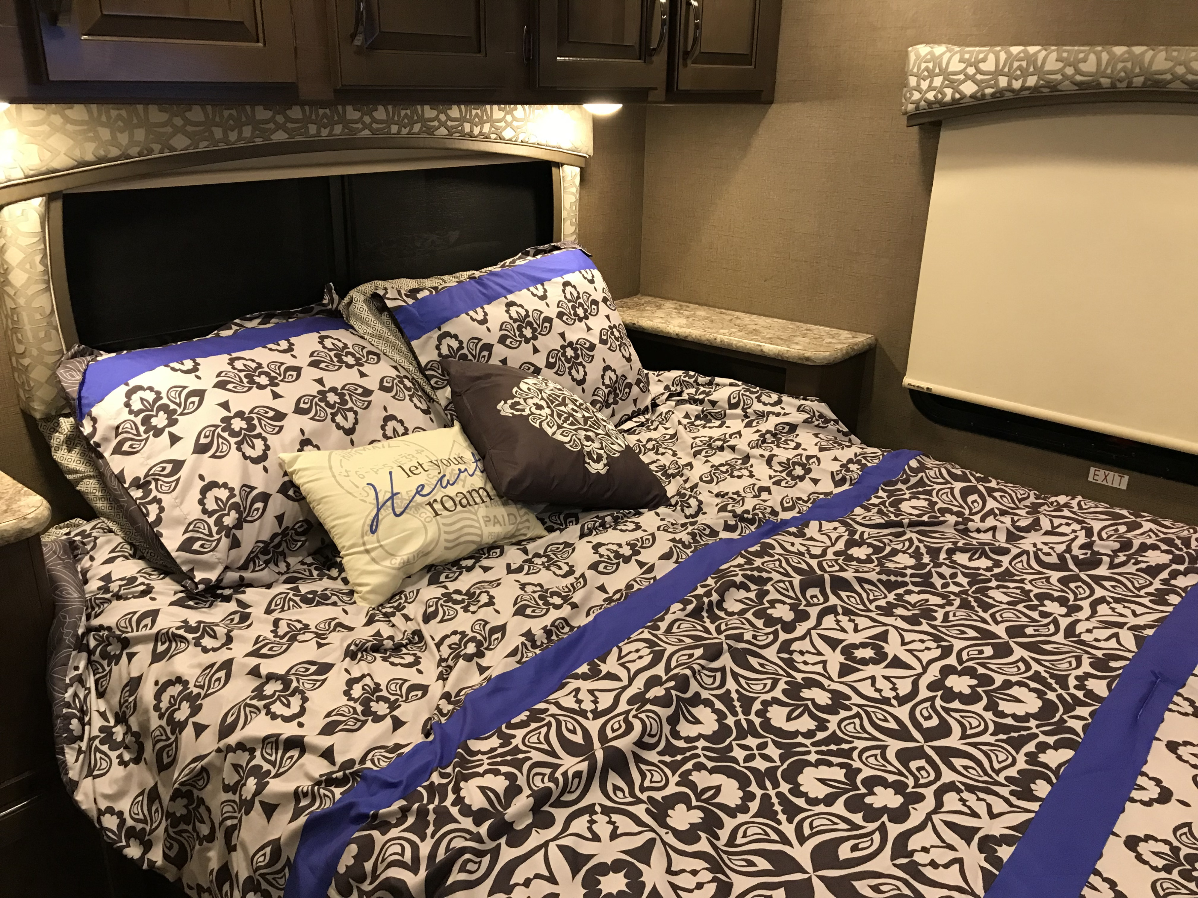 RV rental bed with intricate brown and white patterned bedding, blue accents, overhead cabinets, and a window shade.. Thor Chateau 2020