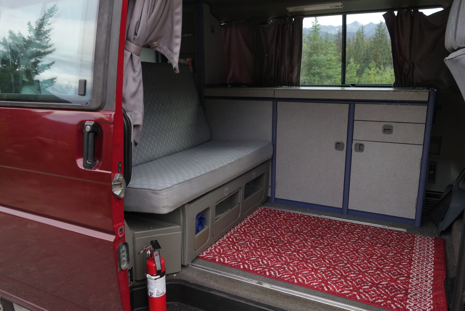 Open RV rental door reveals a camper van interior with a grey bench seat, light cabinets, red rug, and forest mountain views.. Volkswagen Westfalia 1992