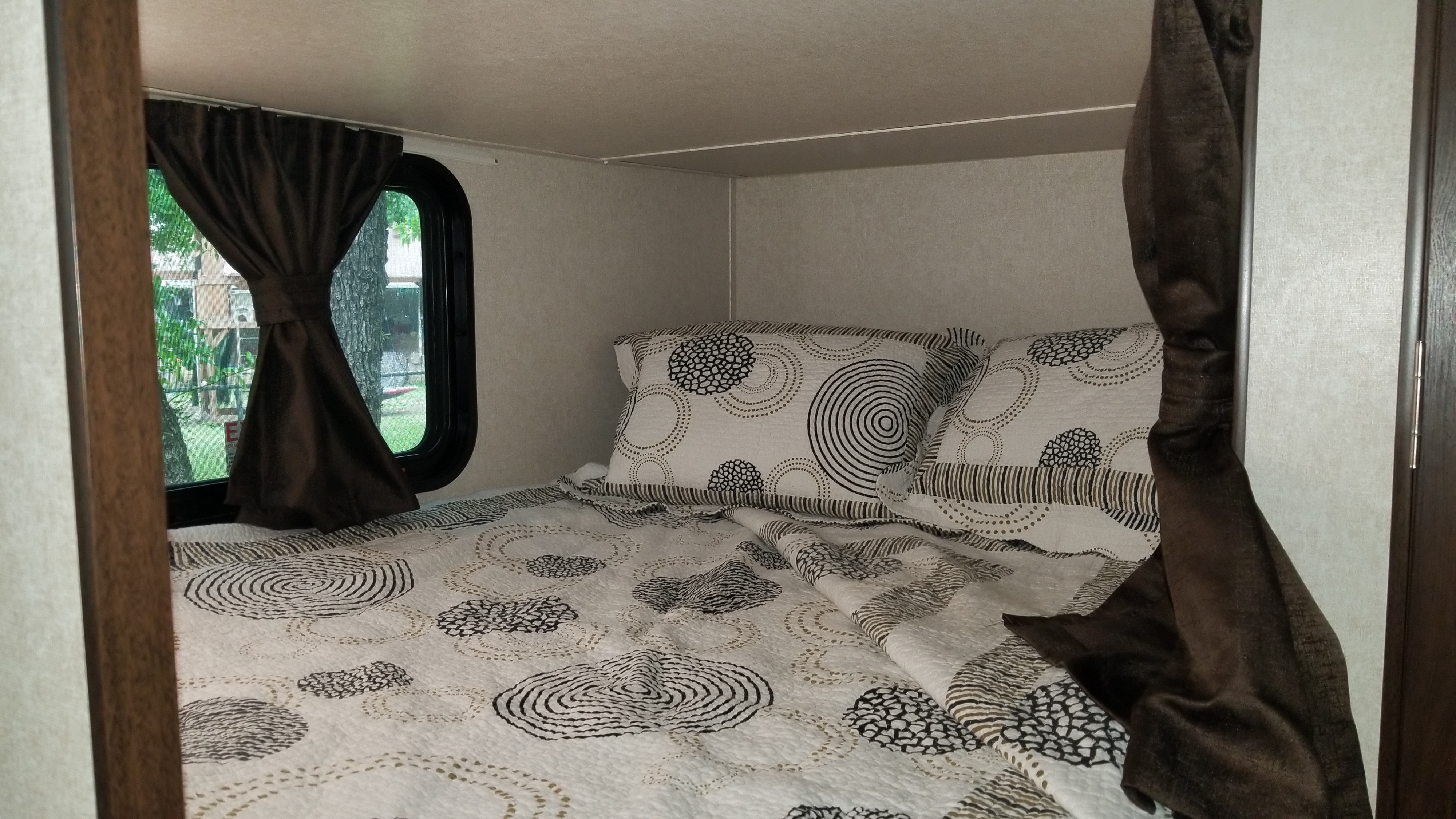 Inside an RV rental: a comfy bed with a white geometric patterned quilt & pillows. Window offers a glimpse of nature.. Jayco Jay Flight 2018