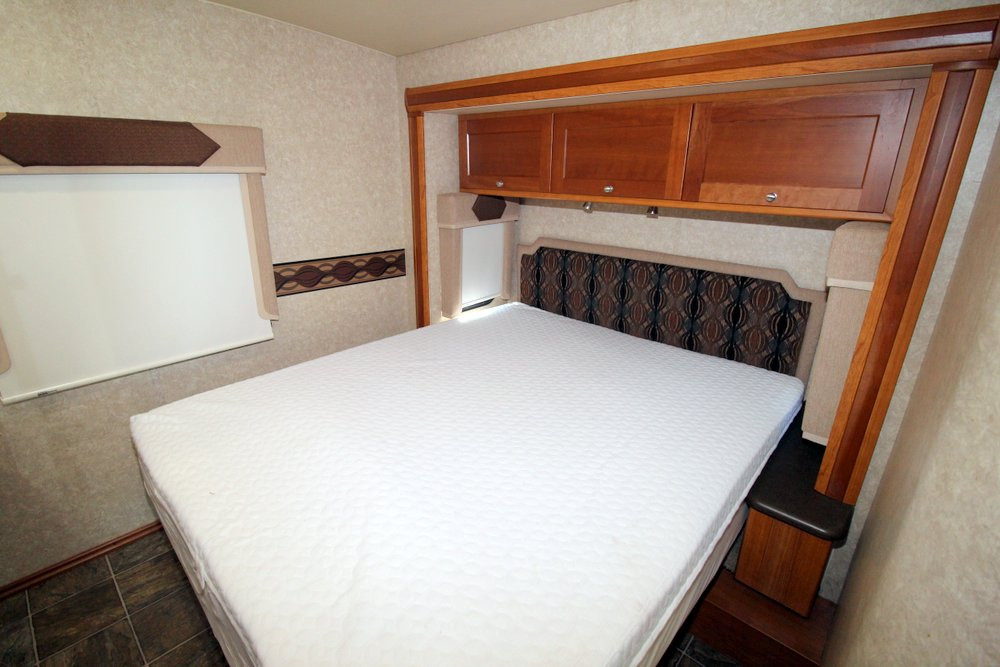 Inside an RV rental: a cozy bedroom with a large, white-mattressed bed, overhead storage, and a window.. Itasca Cambria 2011