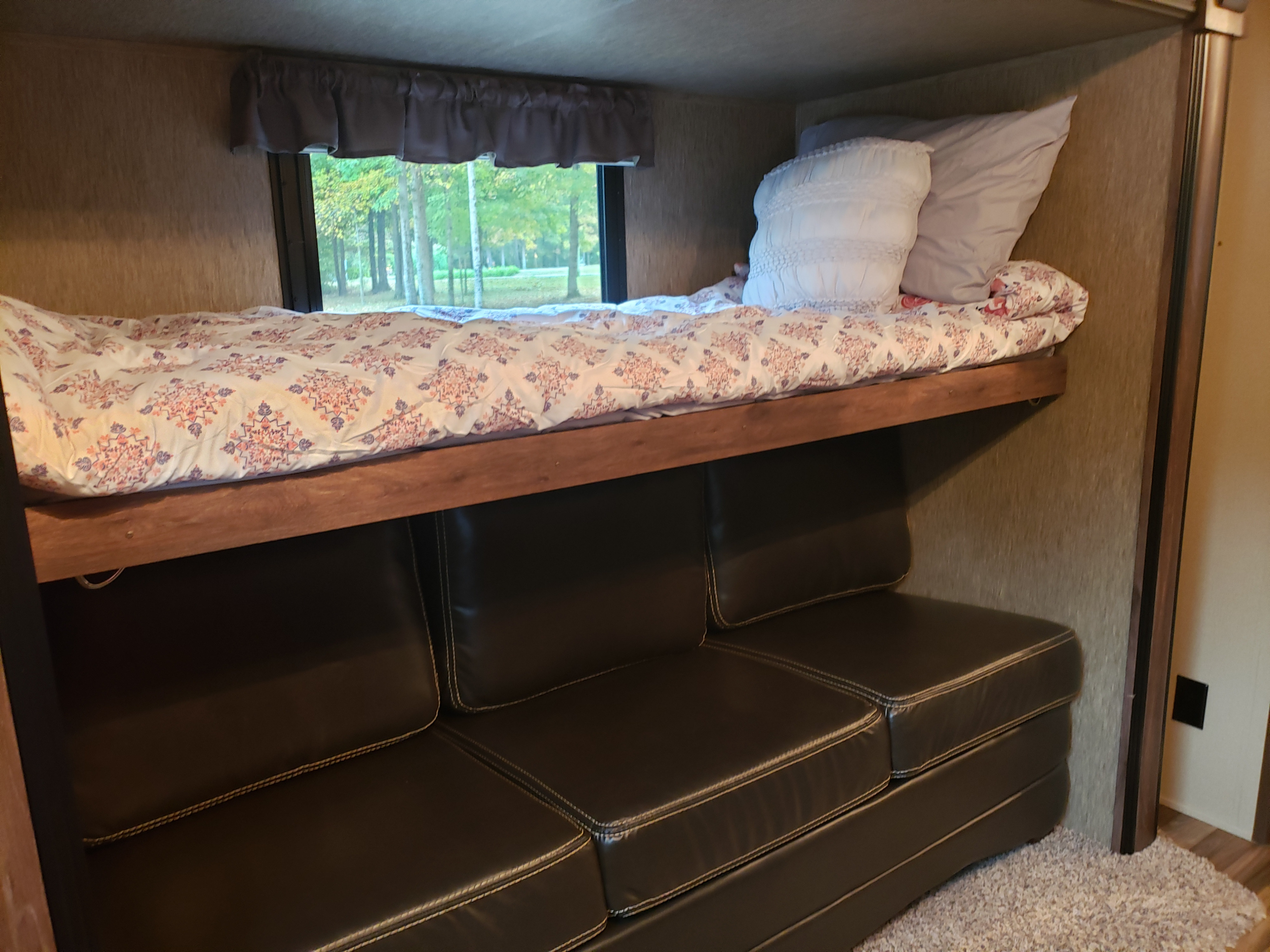 RV rental bunk beds and comfy sofa. The top bunk has a patterned comforter, and a window shows trees outside.. Dutchmen Aerolite 2016