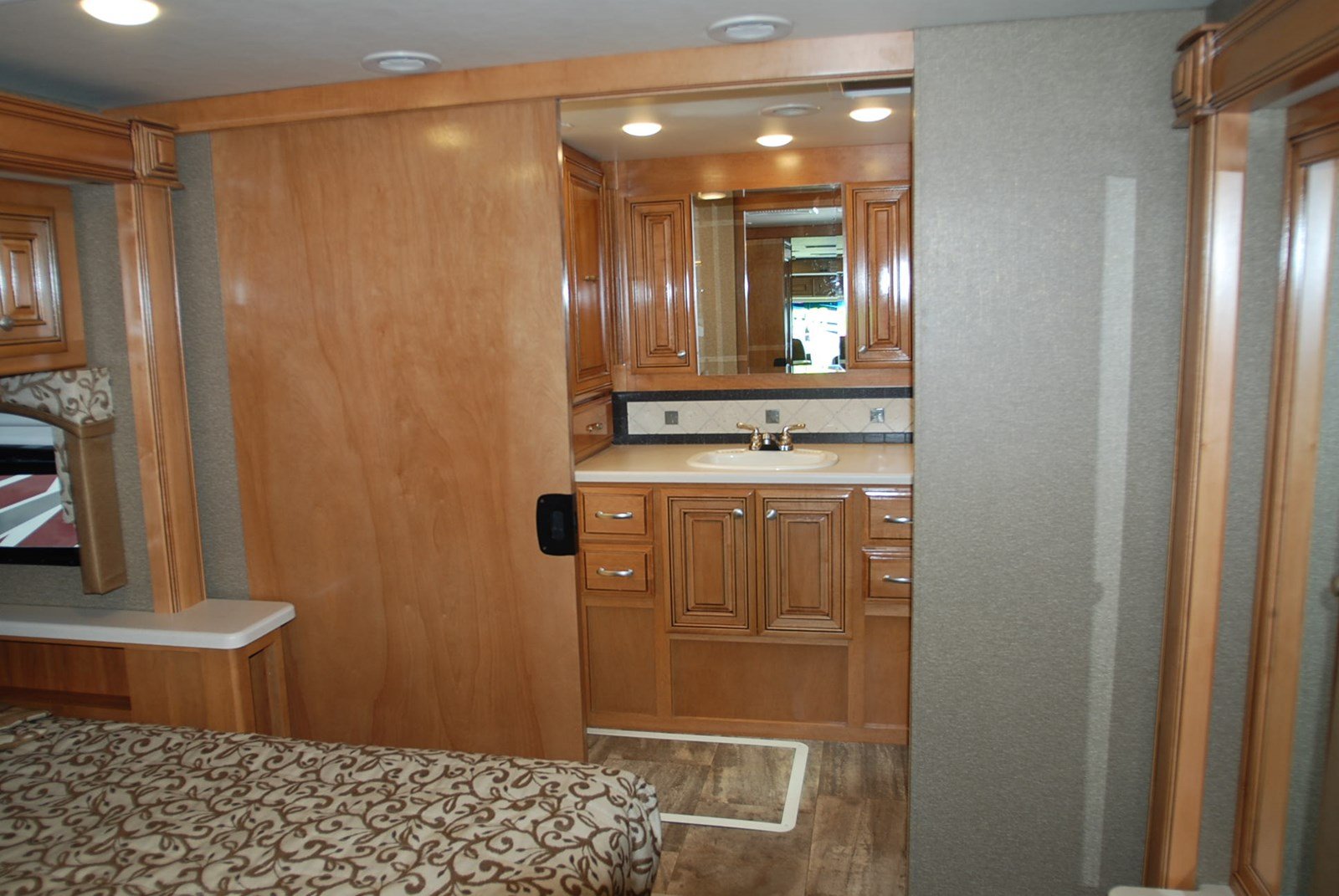 An RV rental interior showing a wooden sliding door open to reveal a bathroom vanity with a sink and mirror, adjacent to a bed.. Thor Palazzo 2016