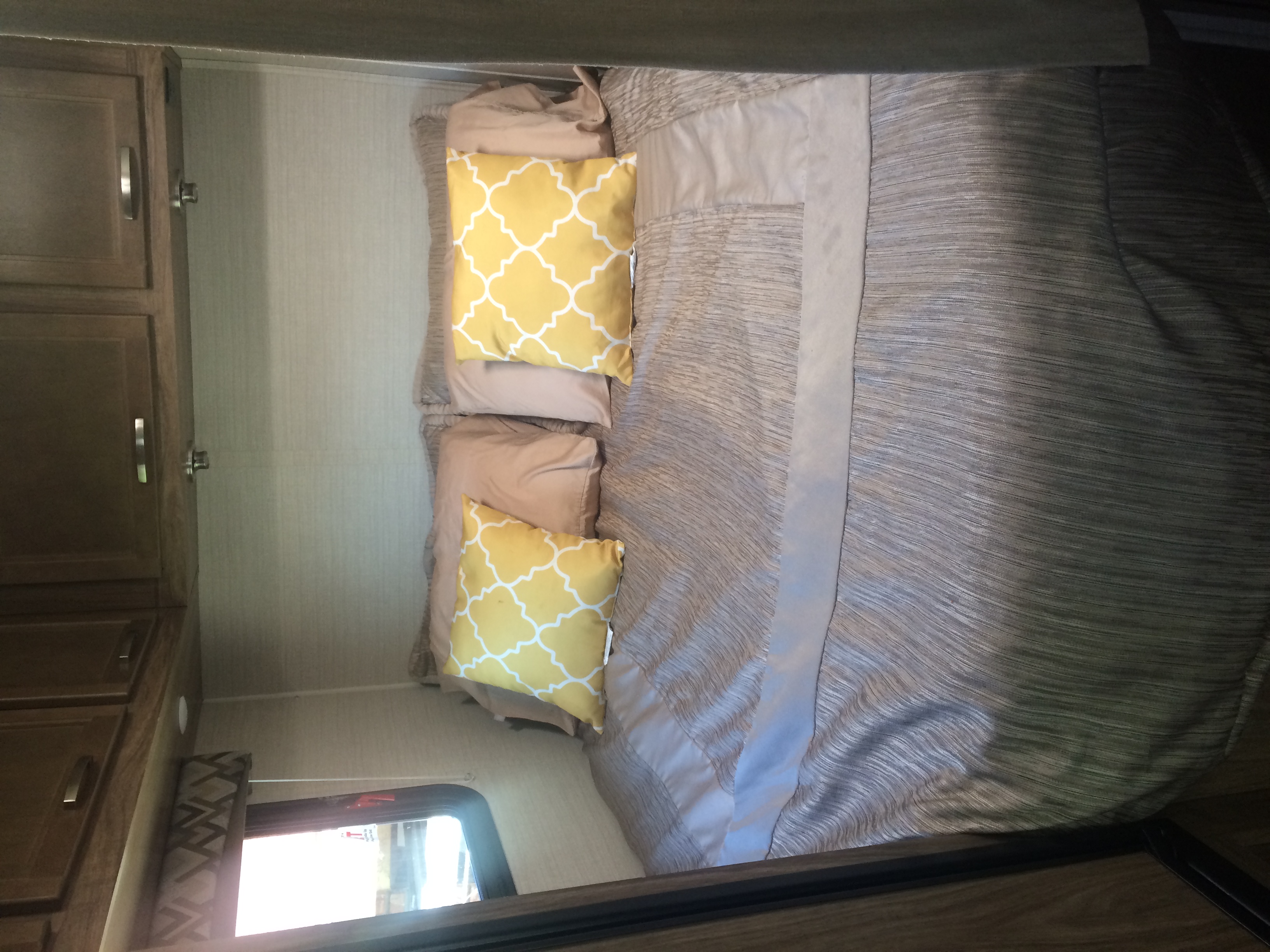 Inside an RV rental: a neat bedroom with striped bedding, yellow geometric pillows, wood cabinetry, and a window.. Jayco Redhawk 2018