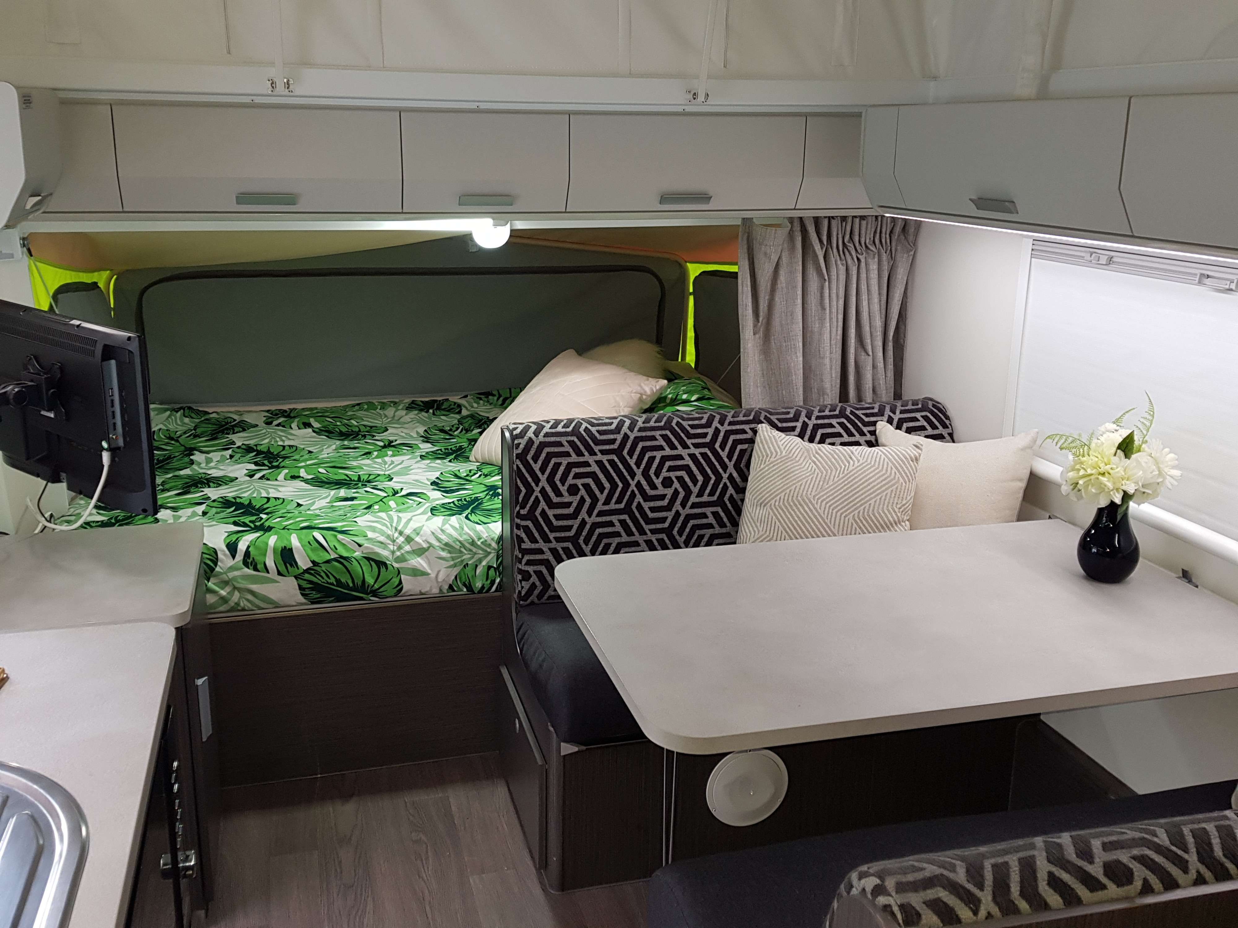 Spacious RV rental interior featuring a cozy bed with leaf print, dining area with modern seating, overhead storage, and a small kitchen setup.. Expanda Outback 2018