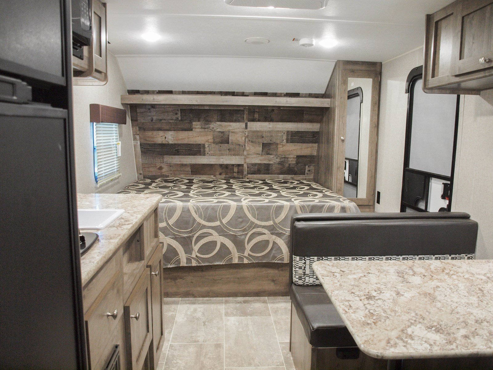 This RV rental interior features a full bed with a rustic wood headboard, a built-in dinette, and a compact kitchen.. Dutchmen Aspen Trail LE 1950BH 2020