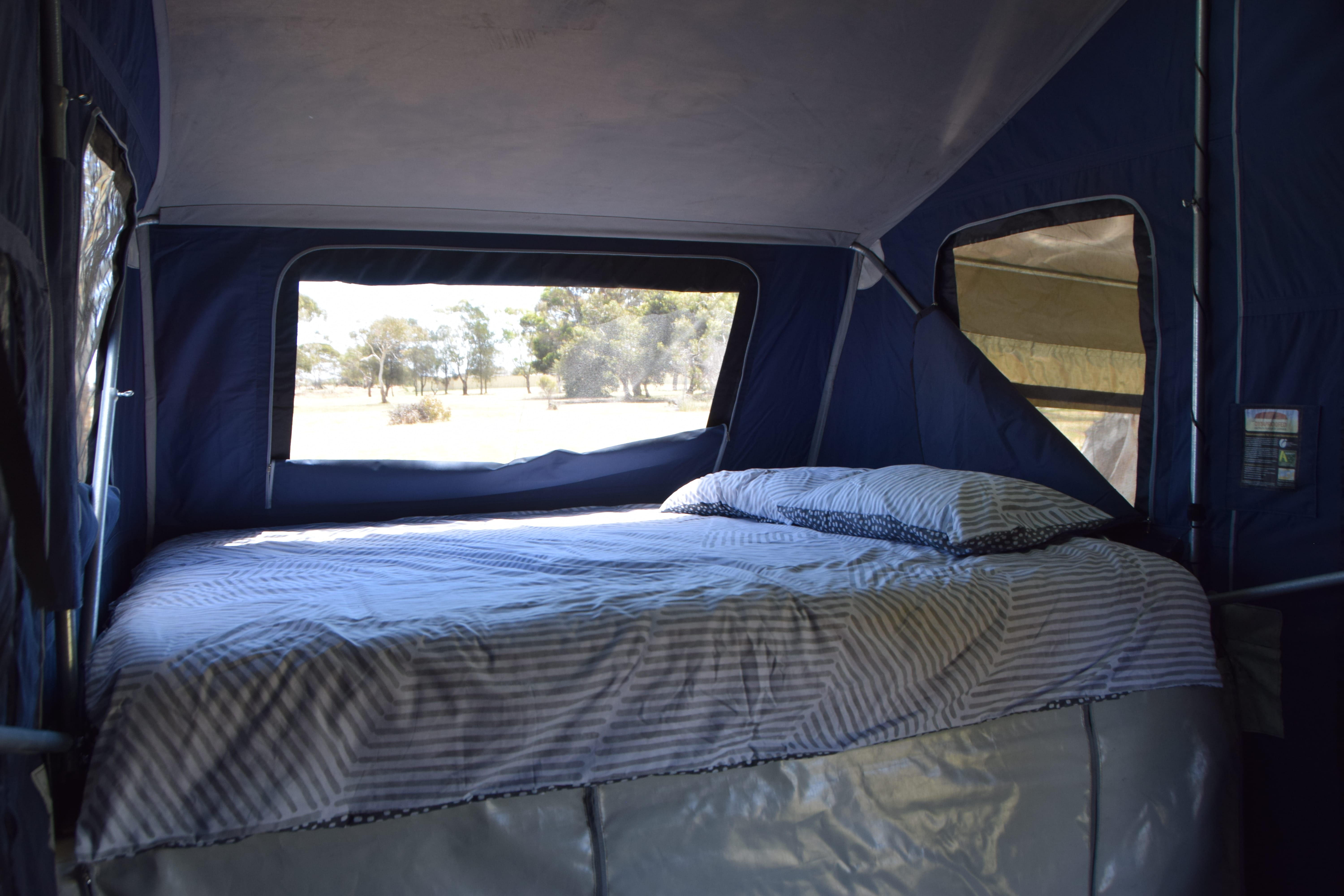 Cozy RV rental bedroom with striped bedding and pillow. Large window reveals a natural outdoor view, perfect for travel and camping.. Johnno's Off-Road 2015