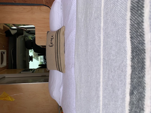 Cozy RV rental interior: bed with white bedding, 'home' pillow, striped blanket, and light wood paneling.. Dodge Sprinter 2500 170ext 2019