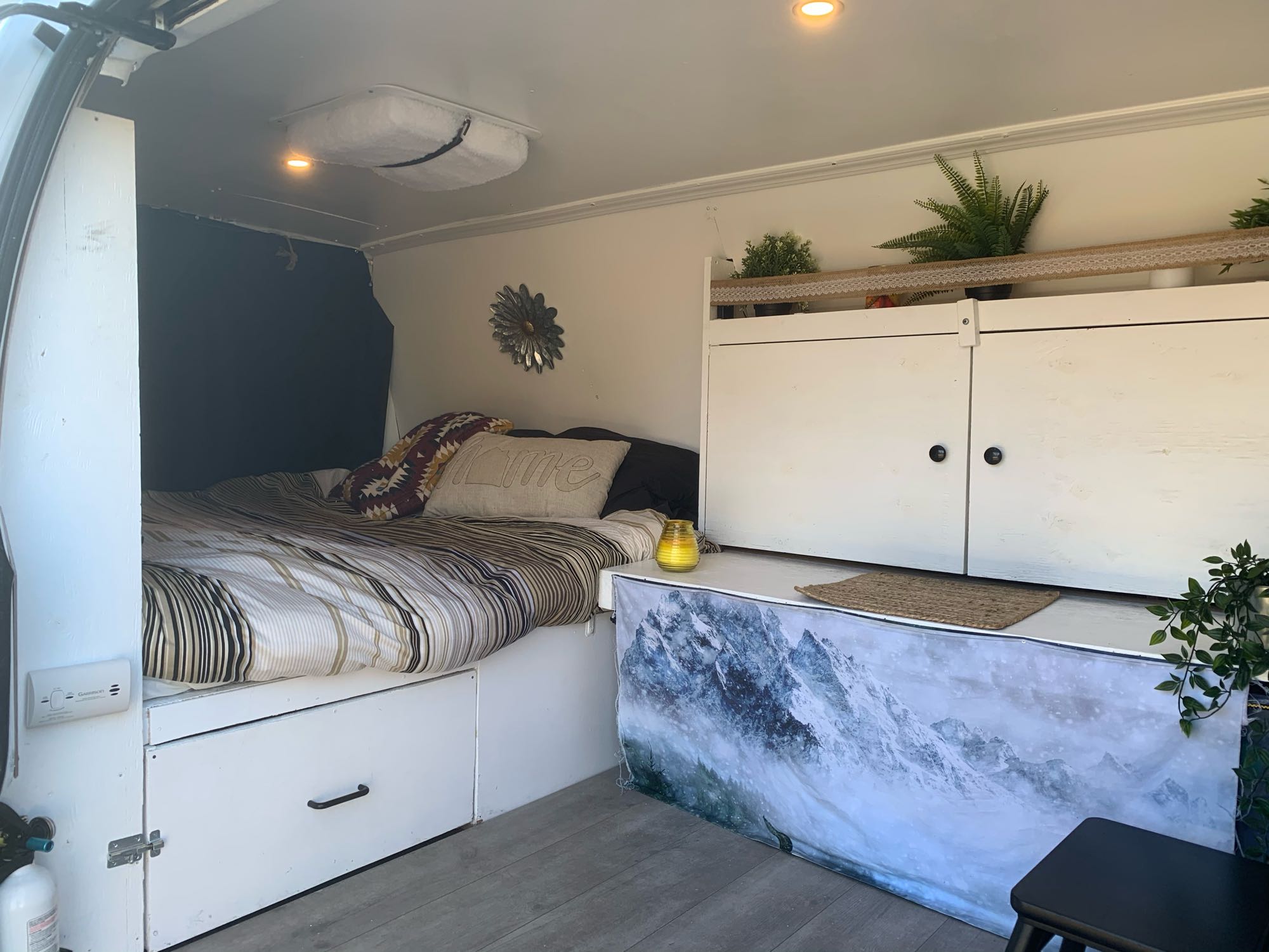 This RV rental interior features a cozy bed with striped linens, white storage cabinets, ceiling lights, and mountain decor.. Chevrolet Express 1500 2006