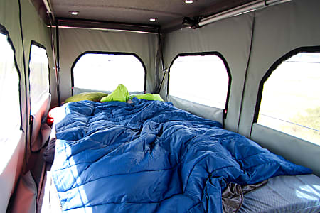 Relax in this RV rental! A comfortable pop-up camper bed with blue bedding and green pillows awaits your adventure.. Dodge Promaster 2018