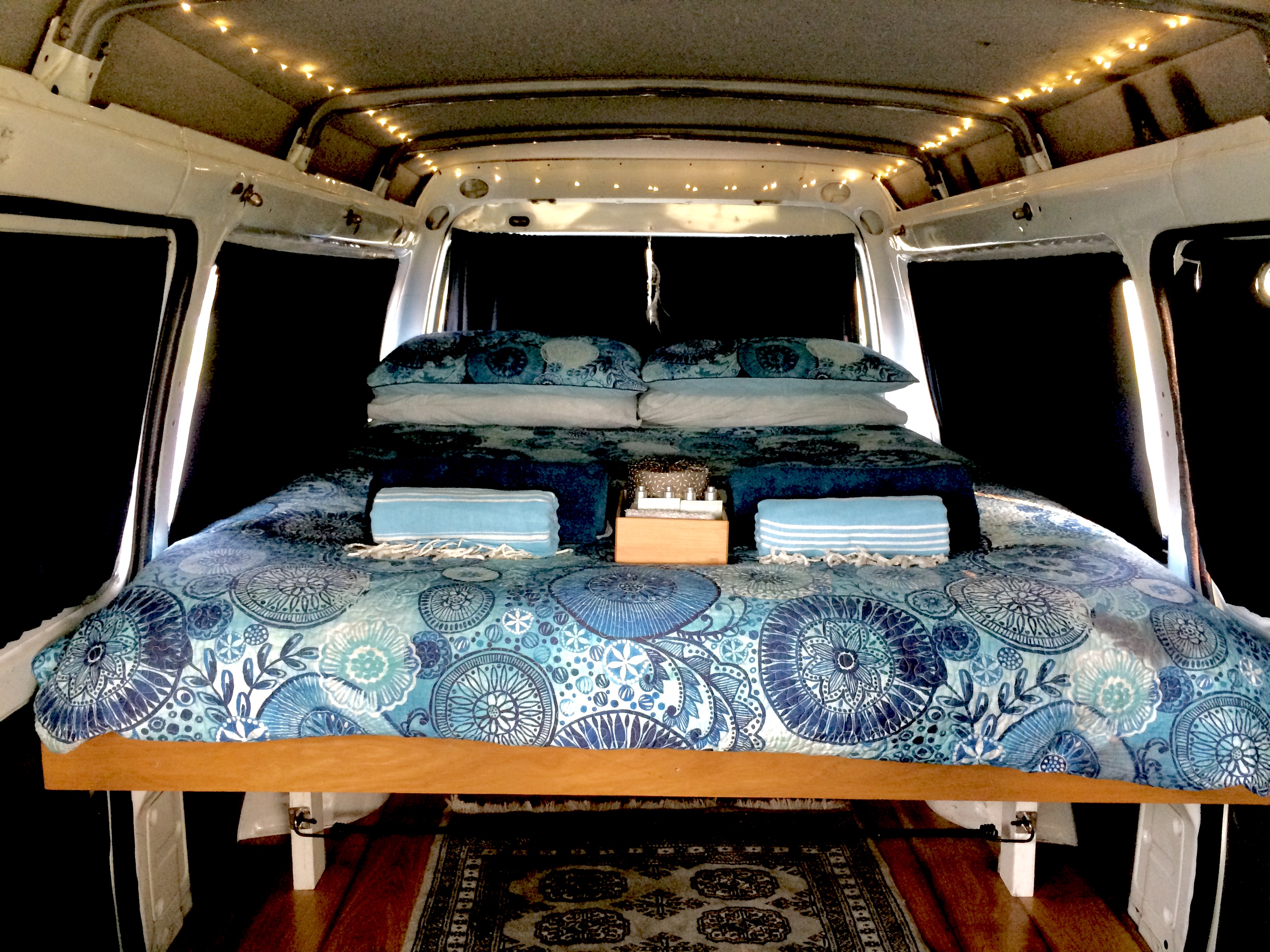 Plush bed inside a modern RV rental, featuring blue quilt, twinkling lights, and blackout curtains for cozy travel.. Mitsubishi Express SJ 1999