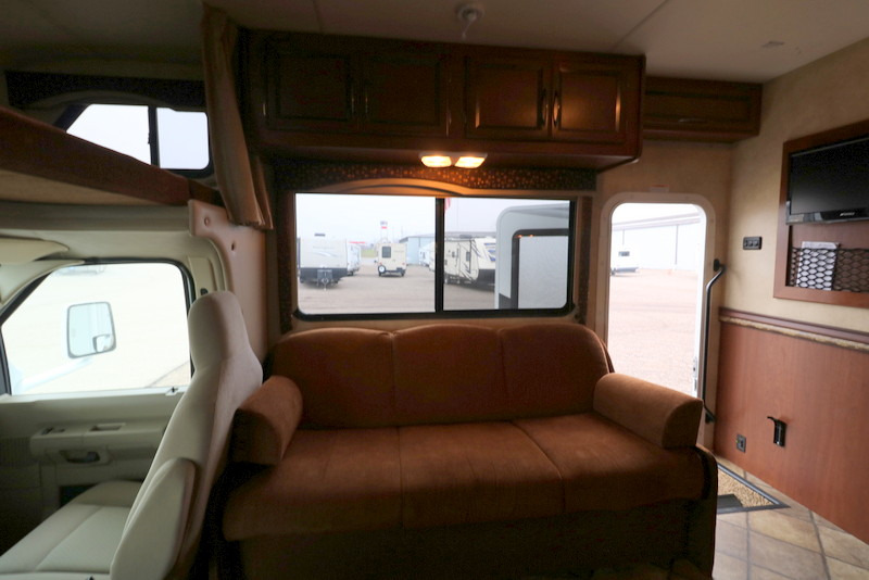RV rental's inviting interior: comfy sofa, overhead bunk, TV, and large windows revealing other RVs.. Thor Motor Coach Freedom Elite 2013