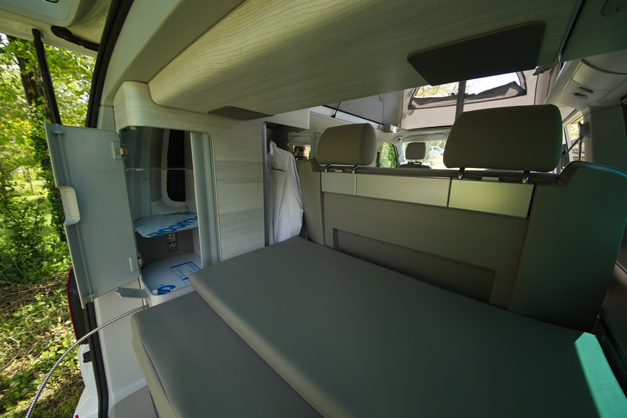 RV rental interior showing a convertible bed, open storage cabinets, and front cabin area. Parked outdoors.. Volkswagen California Coast T6 2021