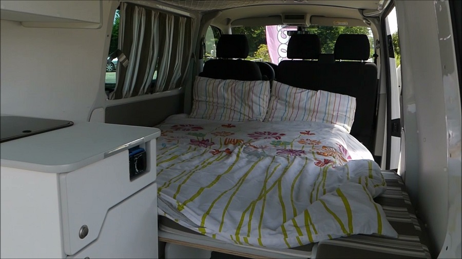 Comfortable RV rental interior with a cozy bed featuring floral and striped bedding, and a compact kitchenette.. Volkswagen Transporter T6 2019