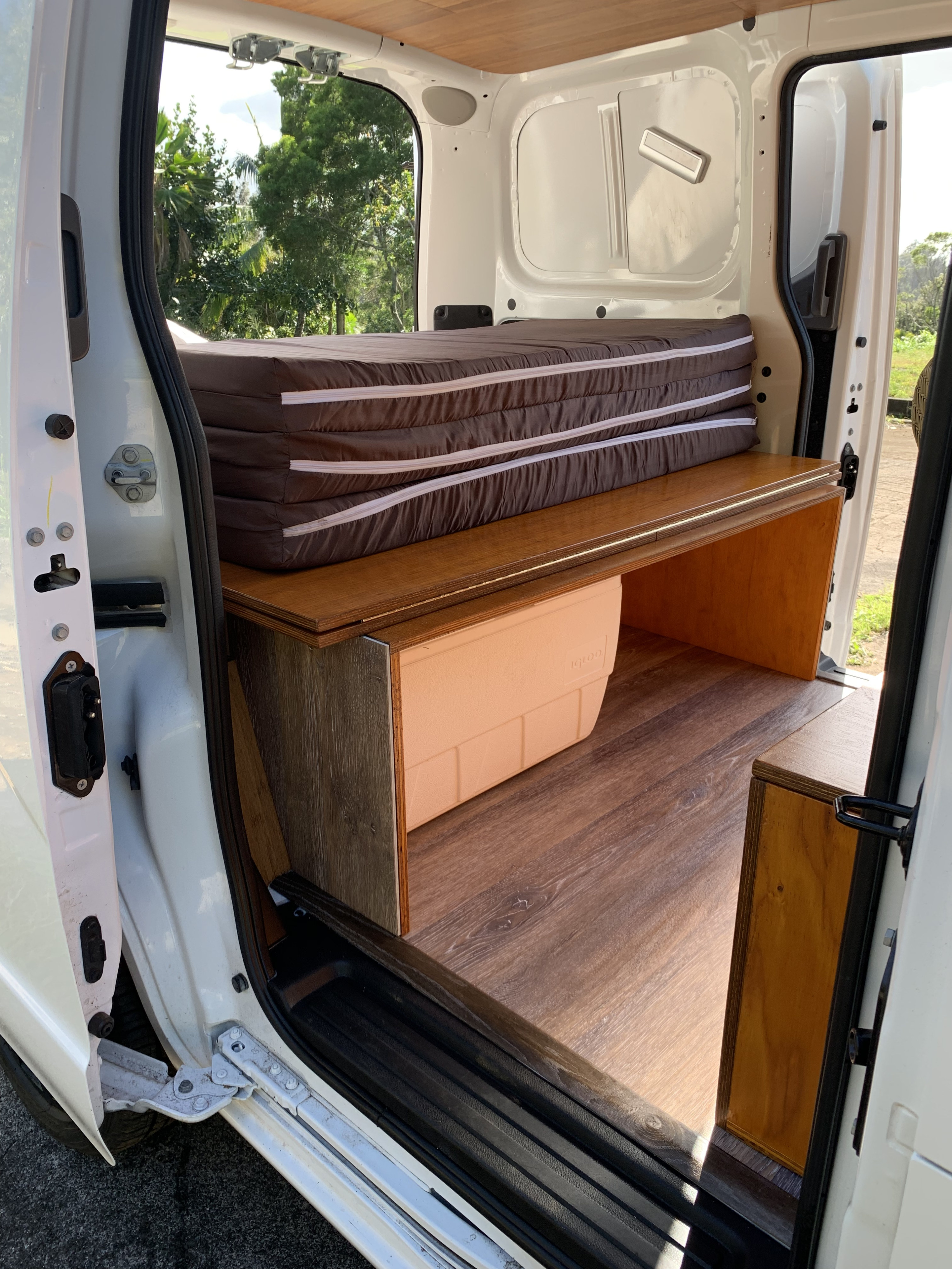 Inside an RV rental camper van: compact bed setup with folded mattress, wood platform, cooler, & storage.. Nissan NV200 2017