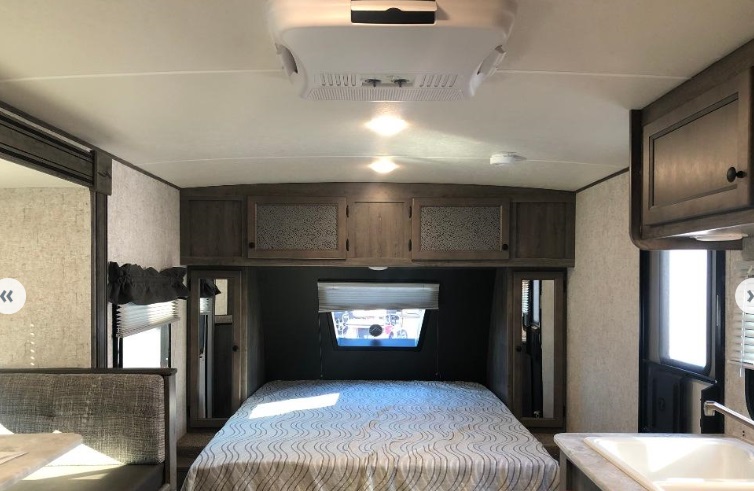 Inside an RV rental: a compact bedroom with bed, overhead storage, and AC unit. Also a kitchen glimpse.. Coachmen Apex 2020