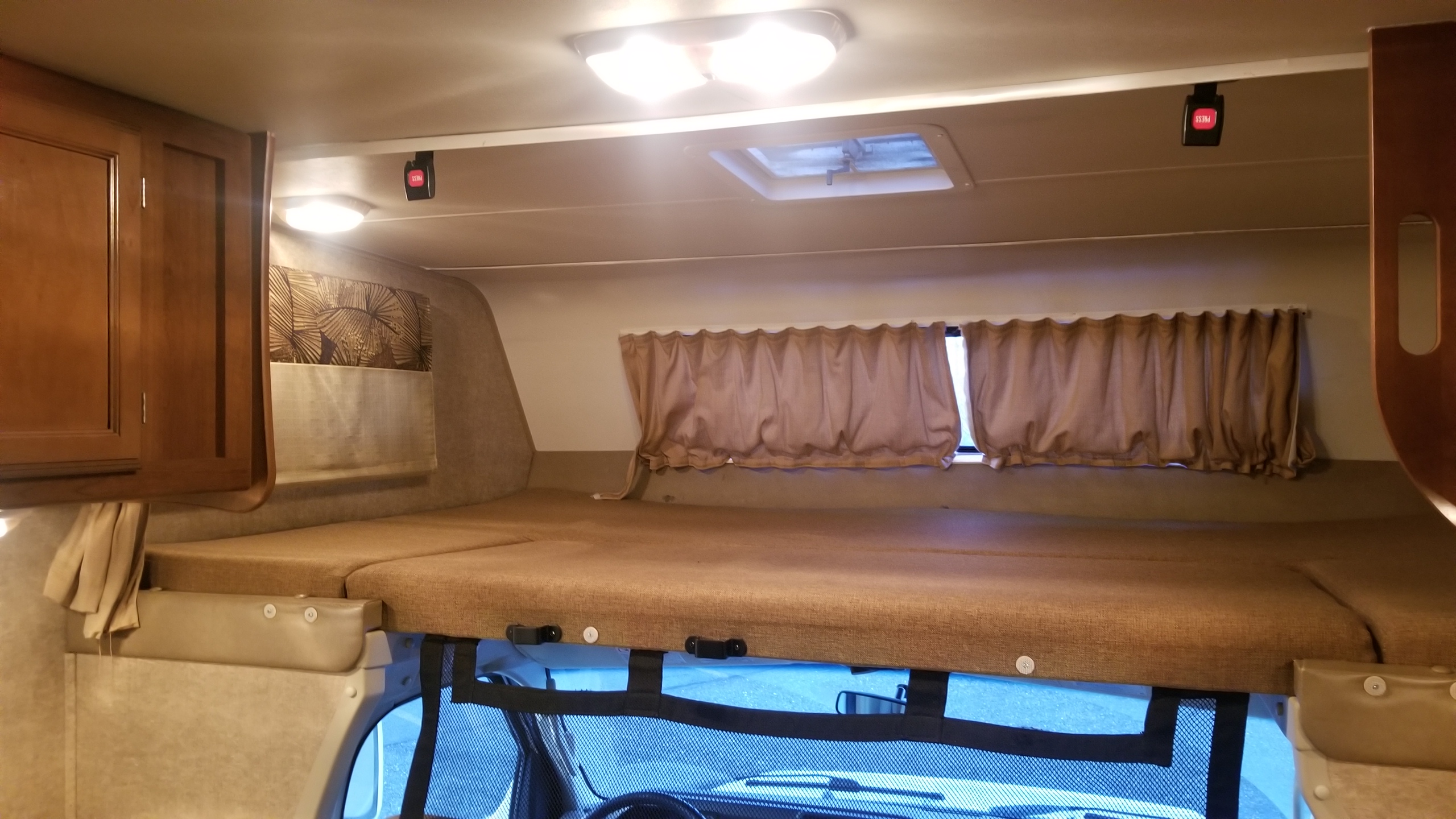 RV rental's cab-over bunk: brown bed, curtains, overhead lights, storage, and ceiling safety buckles for fall protection.. Coachmen Leprechaun 2017