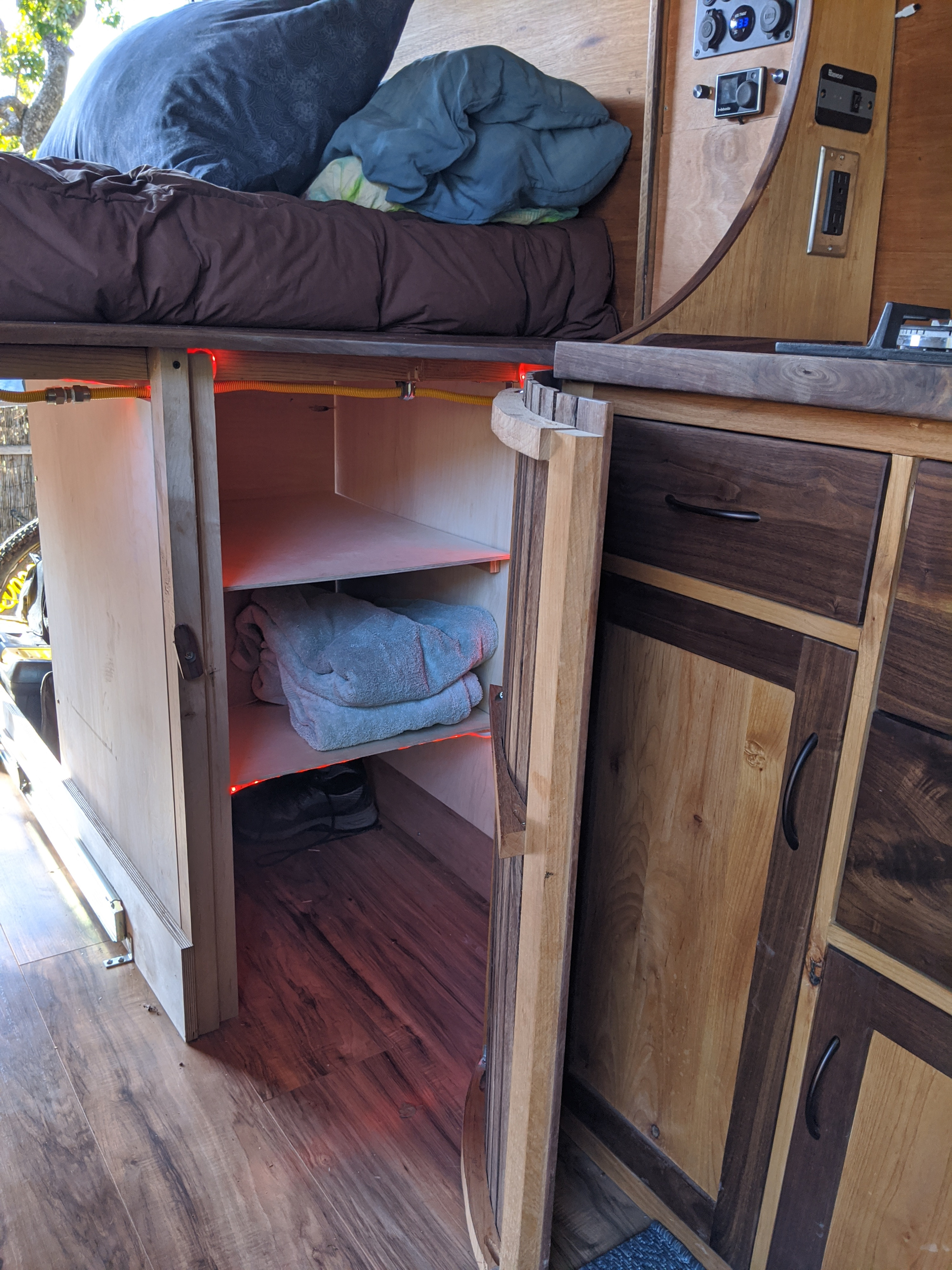Cozy RV rental interior showing a bed, open storage with towels & shoes, and wooden cabinetry. Red lights illuminate shelves.. Mercedes-Benz Sprinter 2019