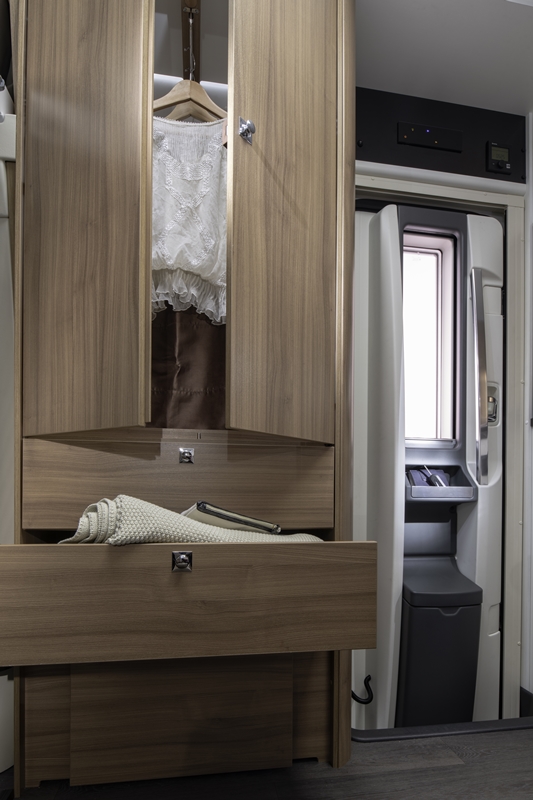 Inside an RV rental: wooden wardrobe with clothes and open drawers, beside a compact entry/utility area with window.. Adria Coral 600 DP 2020