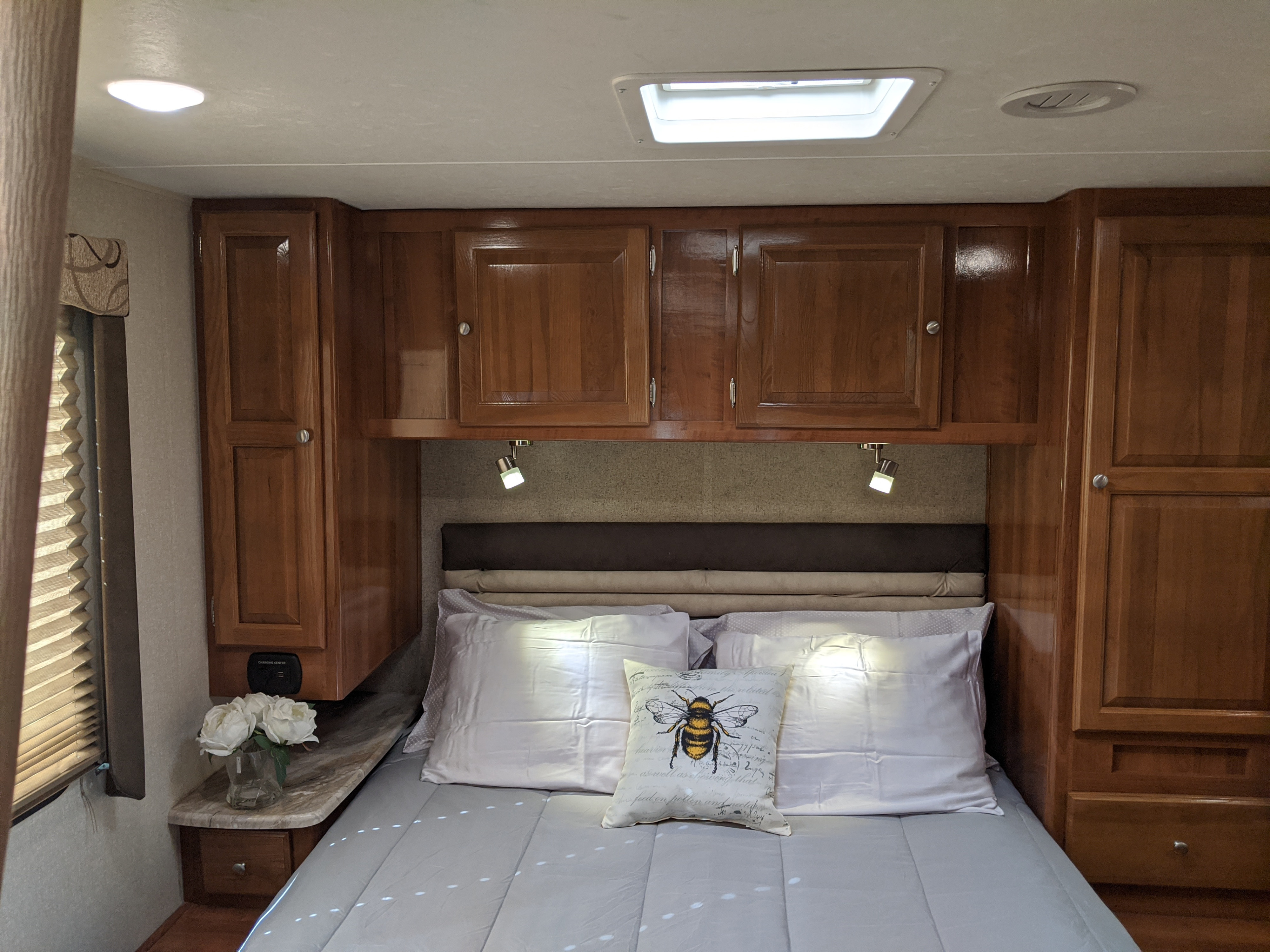 Inviting RV rental bedroom with overhead wood cabinets, cozy bedding, charging station, and reading lights, ready for your trip.. Coachmen Leprechaun 2019