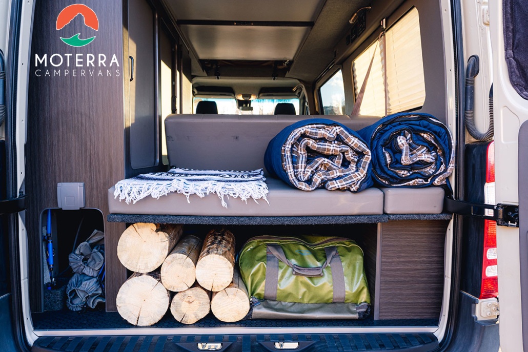 Inside a Moterra Campervans RV rental, showing cozy bedding, stacked firewood, and gear storage for comfortable adventures.. Mercedes-Benz Sprinter 2020