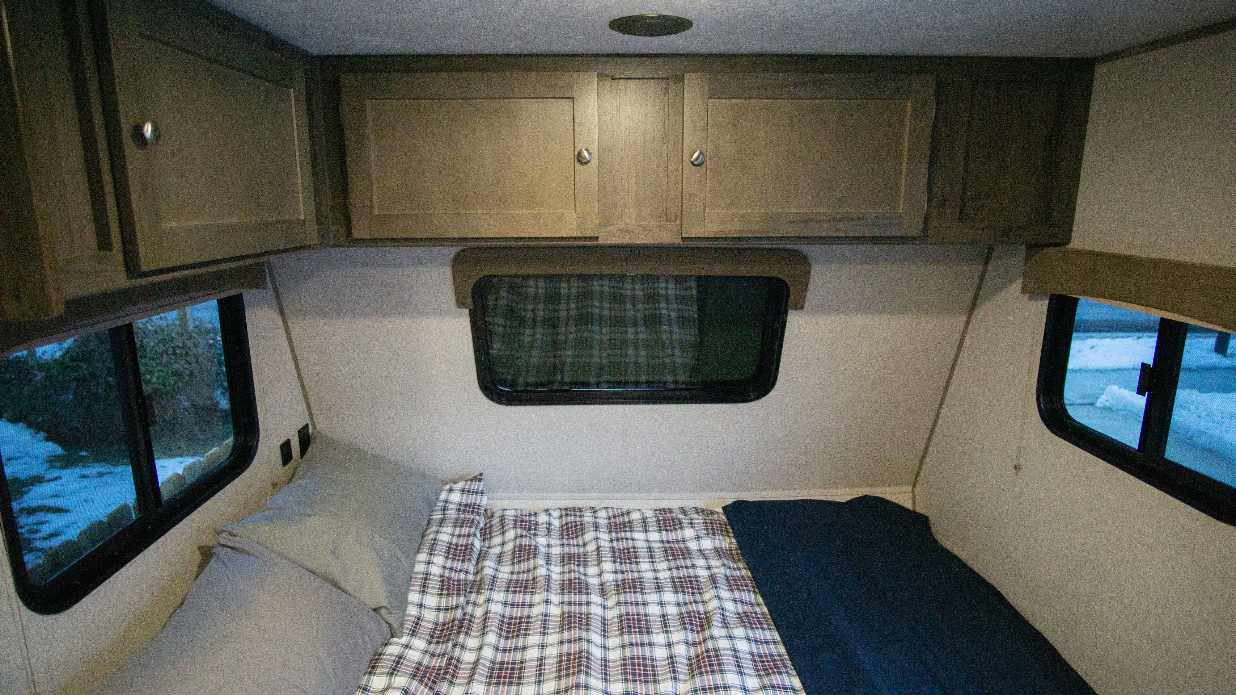 Inside an RV rental: a cozy sleeping area with overhead cabinets, a plaid bed, and windows showing a snowy landscape.. Forest River Clipper Towable 2020