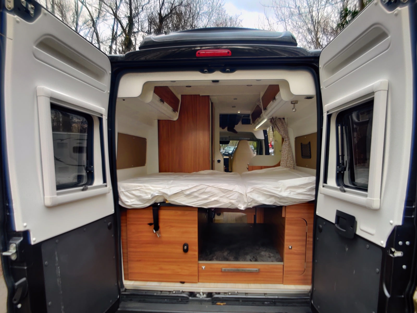 Rear view of an RV rental's interior, featuring a bed, ample storage cabinets, and open back doors for easy loading.. Pilote V600 Toit relevable 2024