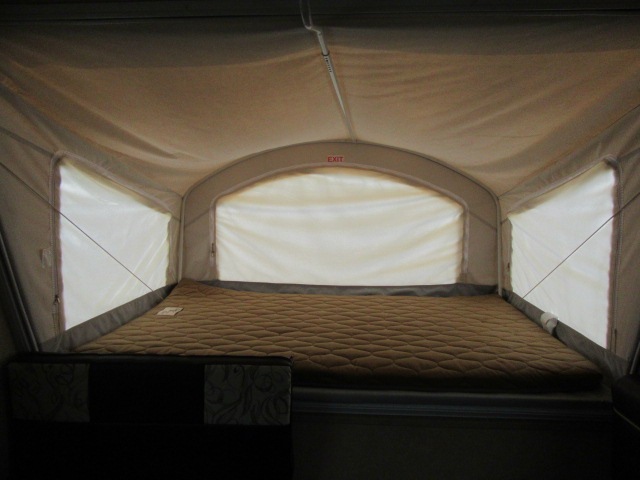 Inside an RV rental pop-up camper: a quilted bed, soft walls, and windows for a cozy sleeping space. "EXIT" sign visible.. Coachmen Catalina 2020