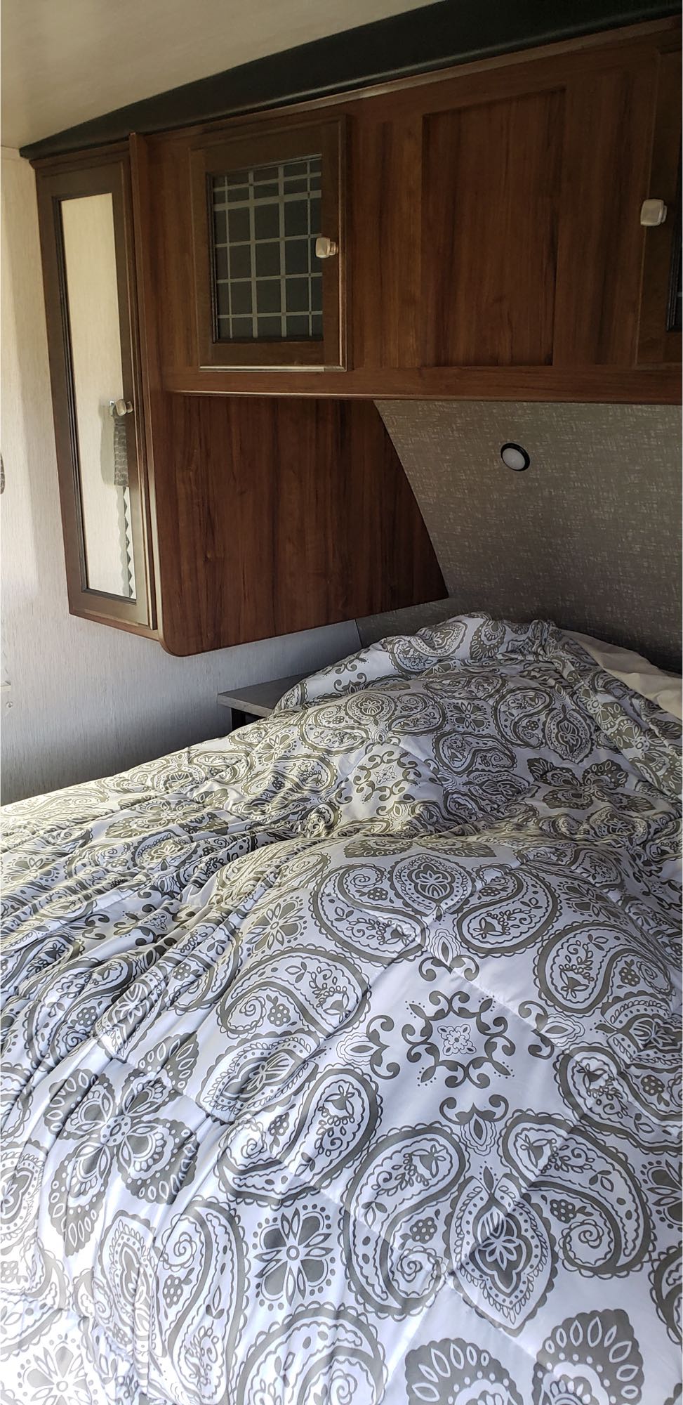 Cozy RV rental bedroom featuring a bed with a white and gray paisley comforter, overhead wooden cabinets, and a mirror.. Heartland M-3385 2019