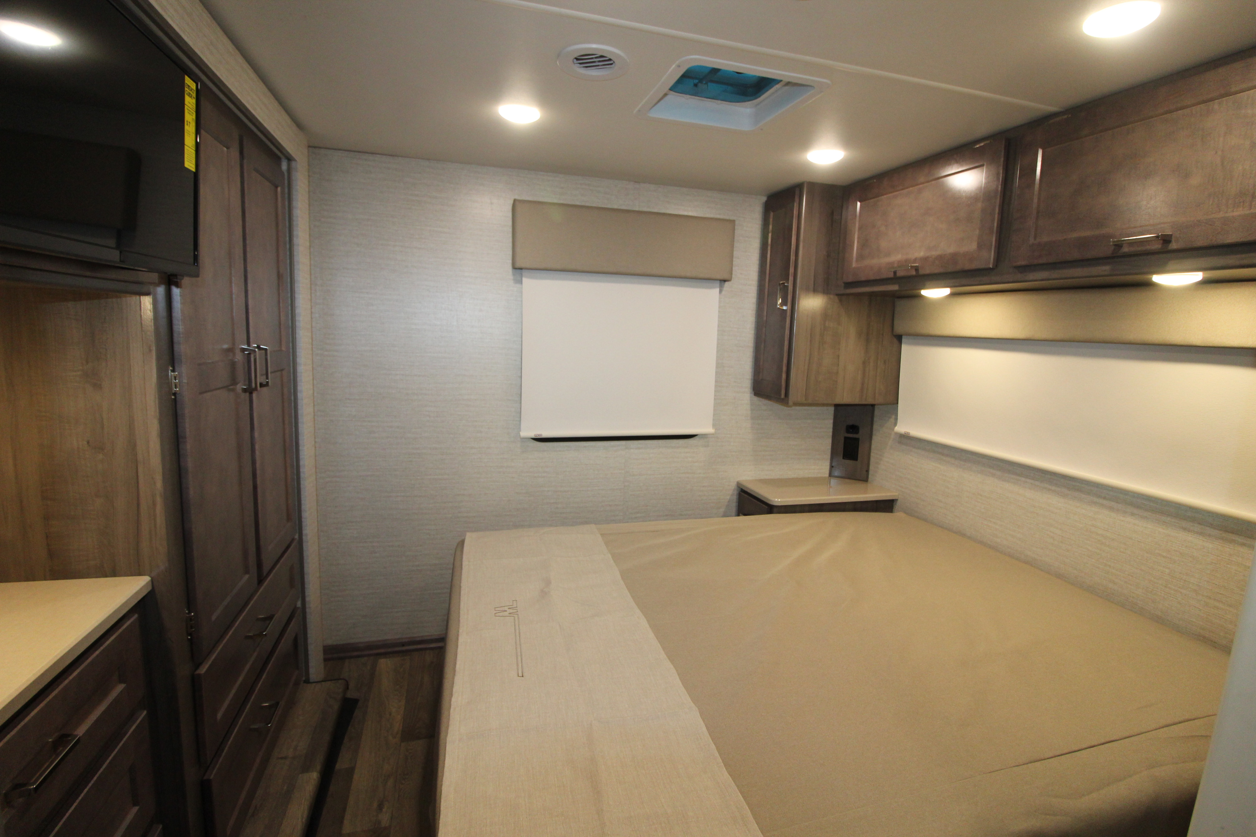 Modern rv rental bedroom with a tan bed, dark wood cabinets and wardrobe, wall-mounted TV, and bright overhead lighting.. Winnebago Minnie Winnie 2020