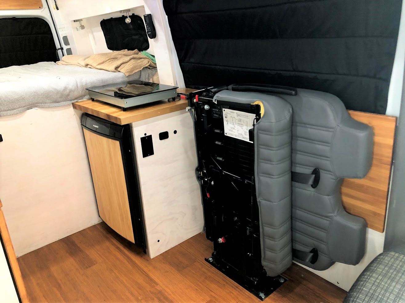 Compact RV rental interior featuring a bed, kitchen with fridge/cooktop, and a foldable seat for versatile living space.. Mercedes-Benz Sprinter 2018
