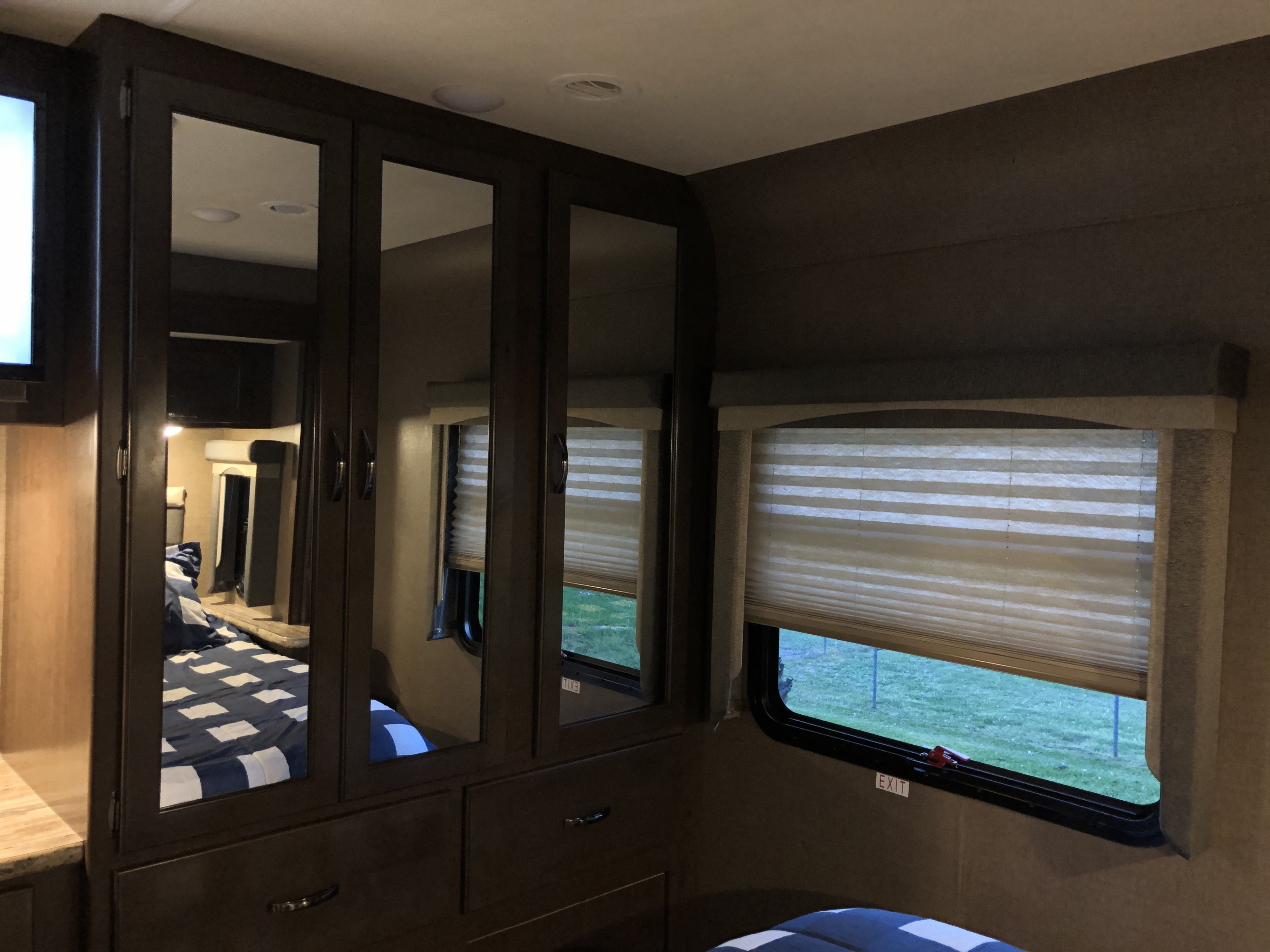 Bedroom in an rv rental: mirrored wardrobe reflects bed with blue/white bedding; window with blinds shows grass & emergency exit.. Thor A.C.E. 2019