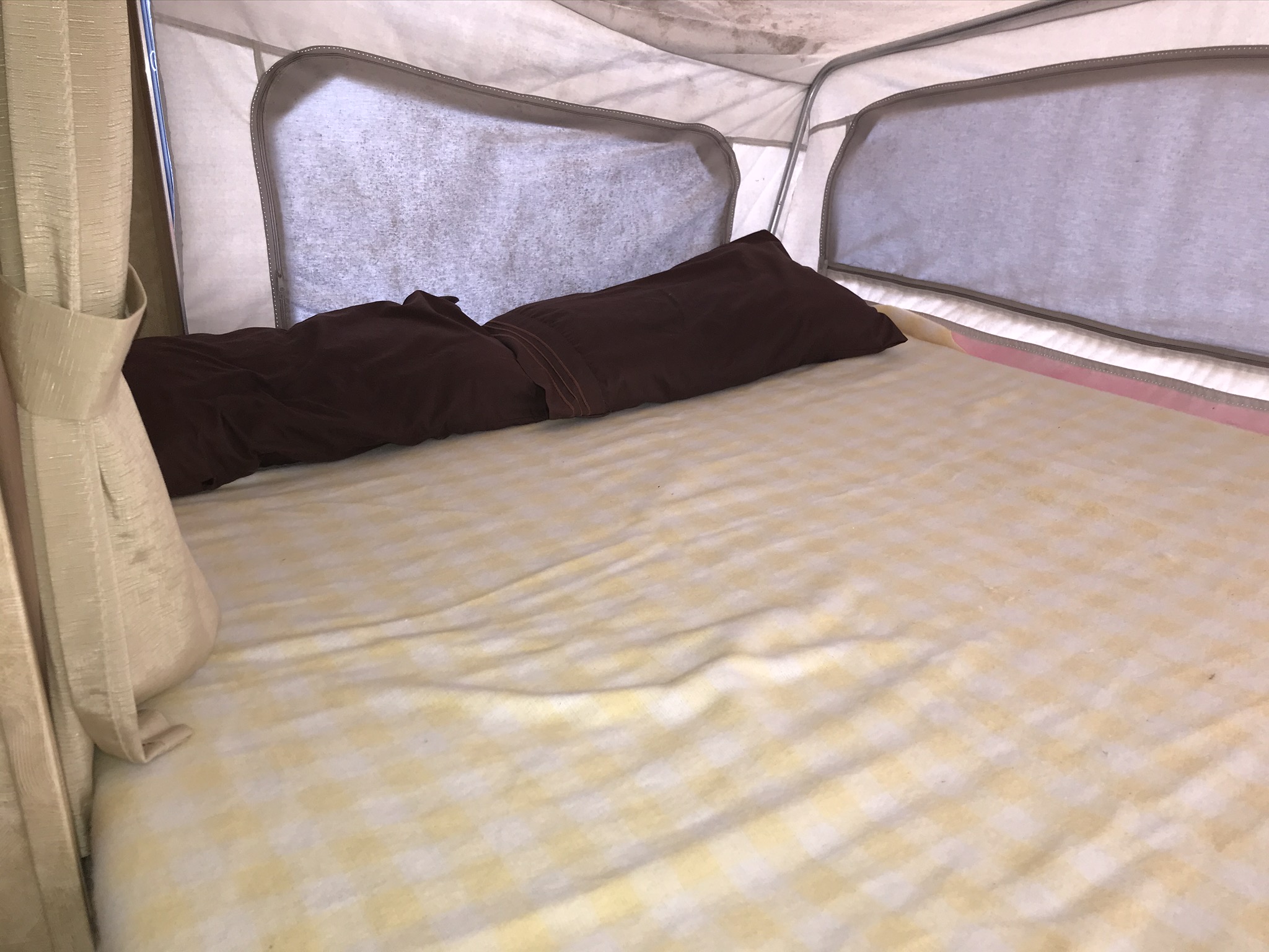 Inside a cozy RV rental pop-up camper: a bed with light checkered sheets, brown pillow, and curtains, ready for your adventure.. Starcraft Antigua 2007