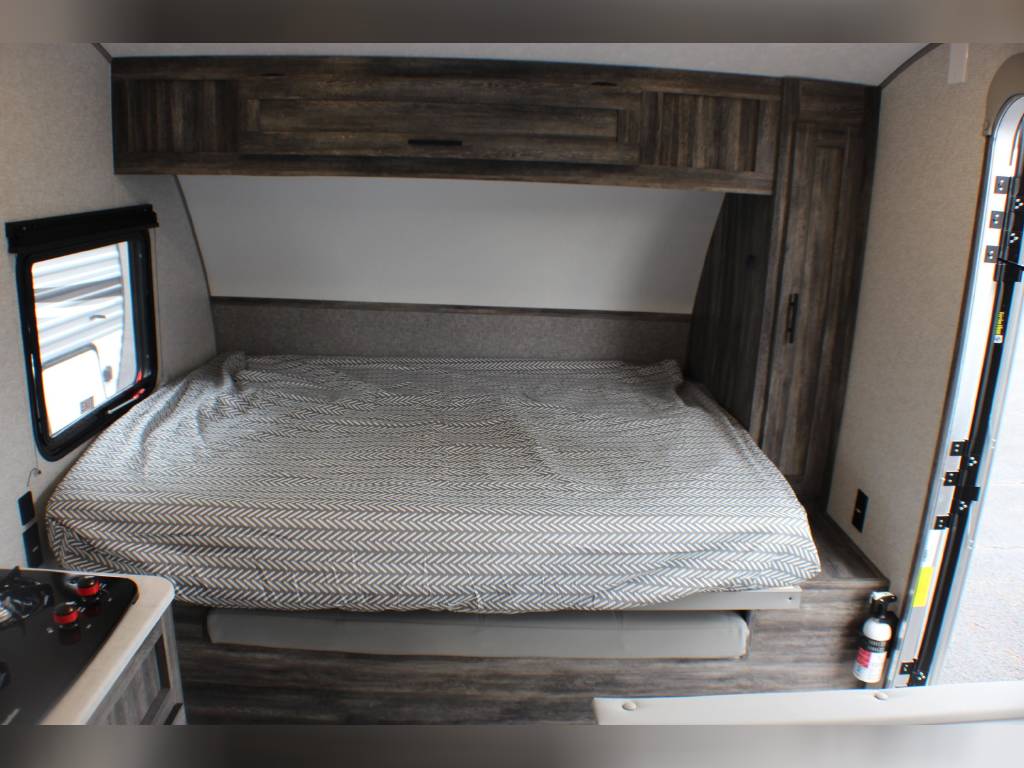 An RV rental bedroom area featuring a large bed, window, overhead cabinets, and a partial view of the kitchen.. Forest River Wildwood FSX 2020