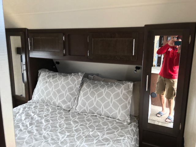 RV rental bedroom featuring a bed with patterned linens, overhead cabinets, and a mirrored wardrobe reflecting a person.. Forest River Cherokee Cascade 2018