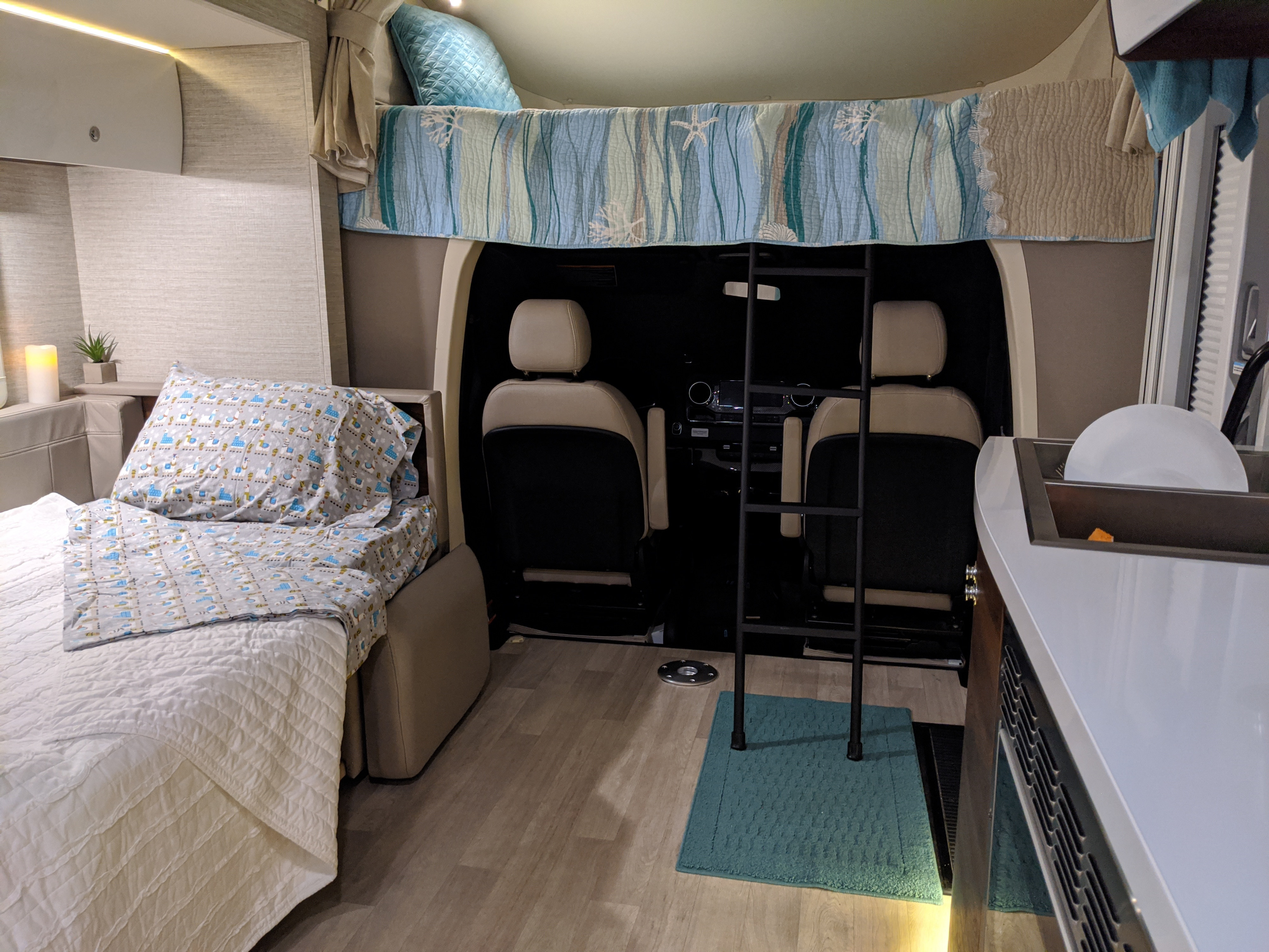 View inside an RV rental showing a comfortable camper van interior with a lower bed, overhead bunk, front cab, and kitchenette.. Winnebago Navion 24D 2020