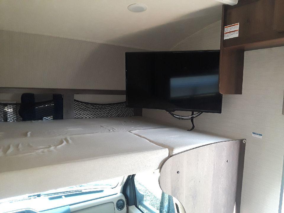 Inside an rv rental: a cozy over-cab bunk bed with a mounted TV and storage nets, ideal for travel comfort.. Jayco Redhawk 2019