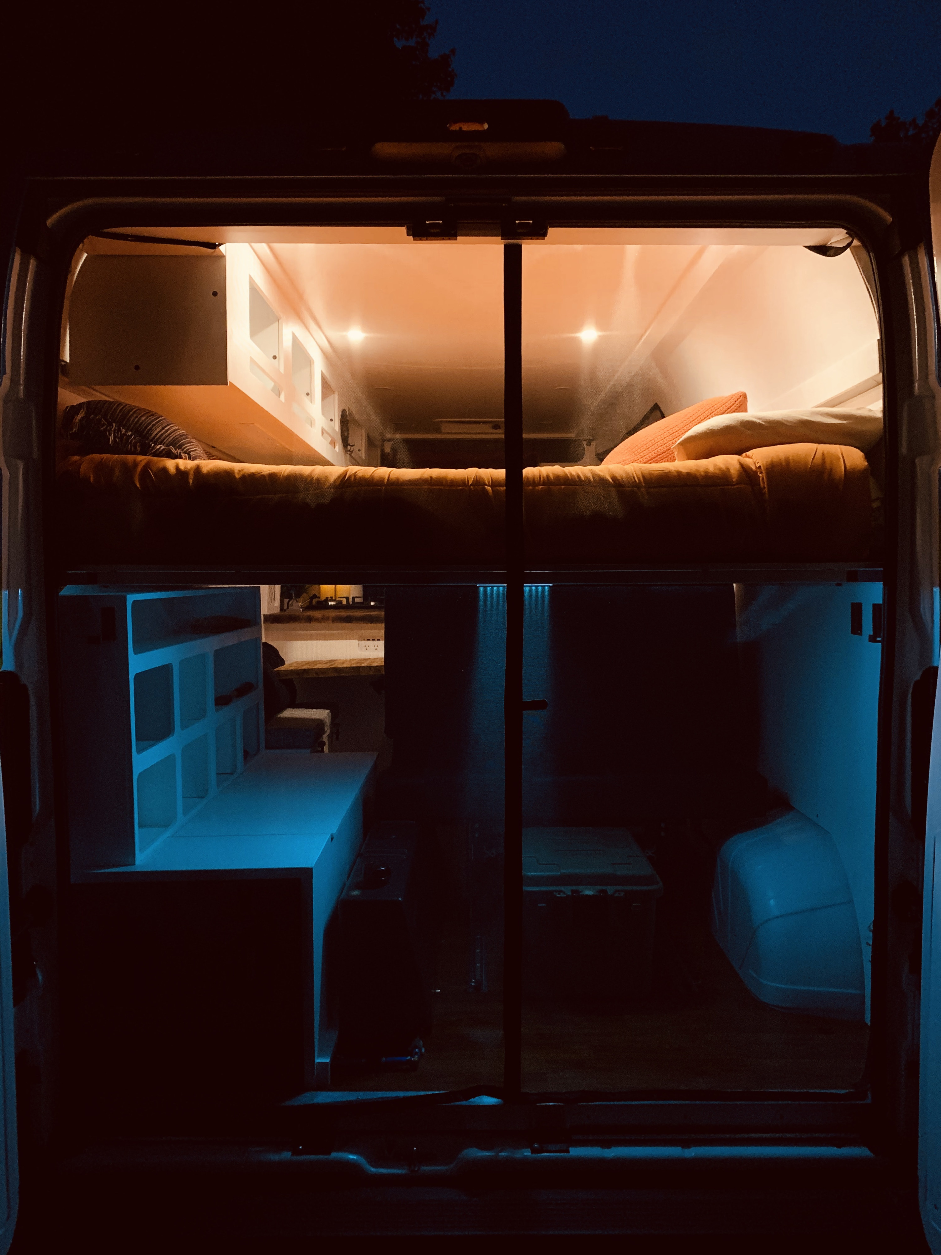 Cozy RV rental interior at night, showing a top bed with warm orange lights and cool blue-lit storage below.. Ram Promaster 2020