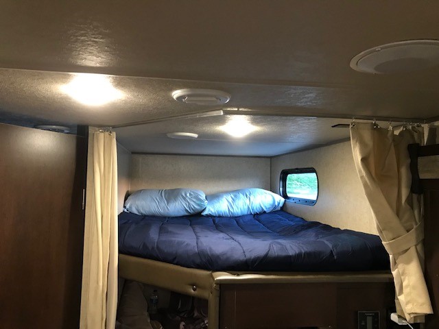 An RV rental's inviting overhead bunk with a blue bed, pillows, window, and privacy curtains.. Forest River Cherokee Cascade 2018