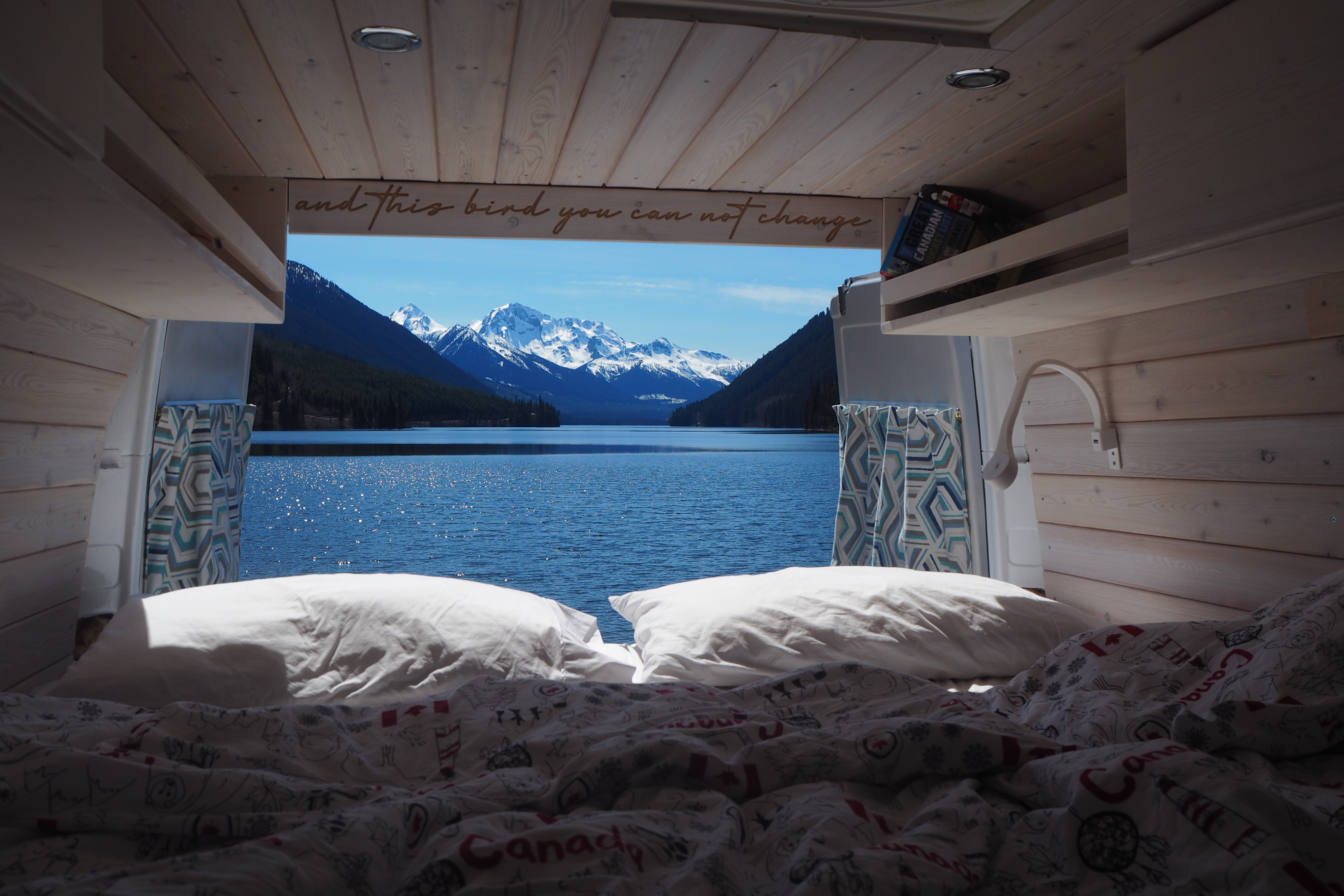 RV rental bed with a stunning view of a serene lake and majestic snow-capped mountains, perfect for a cozy van life adventure.. Mercedes-Benz Sprinter 2002