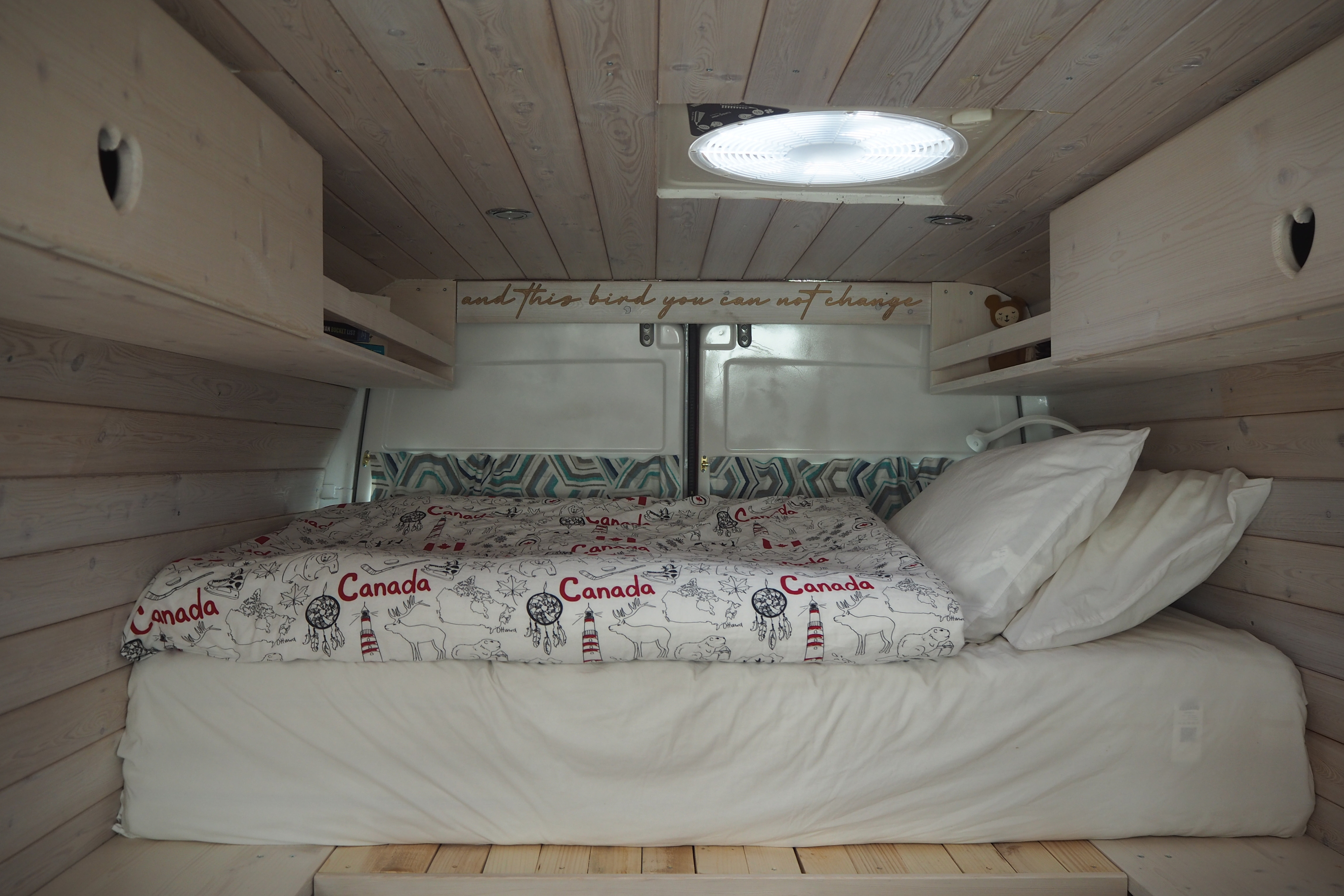 Inside an RV rental: a cozy bed with Canada-themed duvet, white wood paneling, overhead cabinets & light.. Mercedes-Benz Sprinter 2002